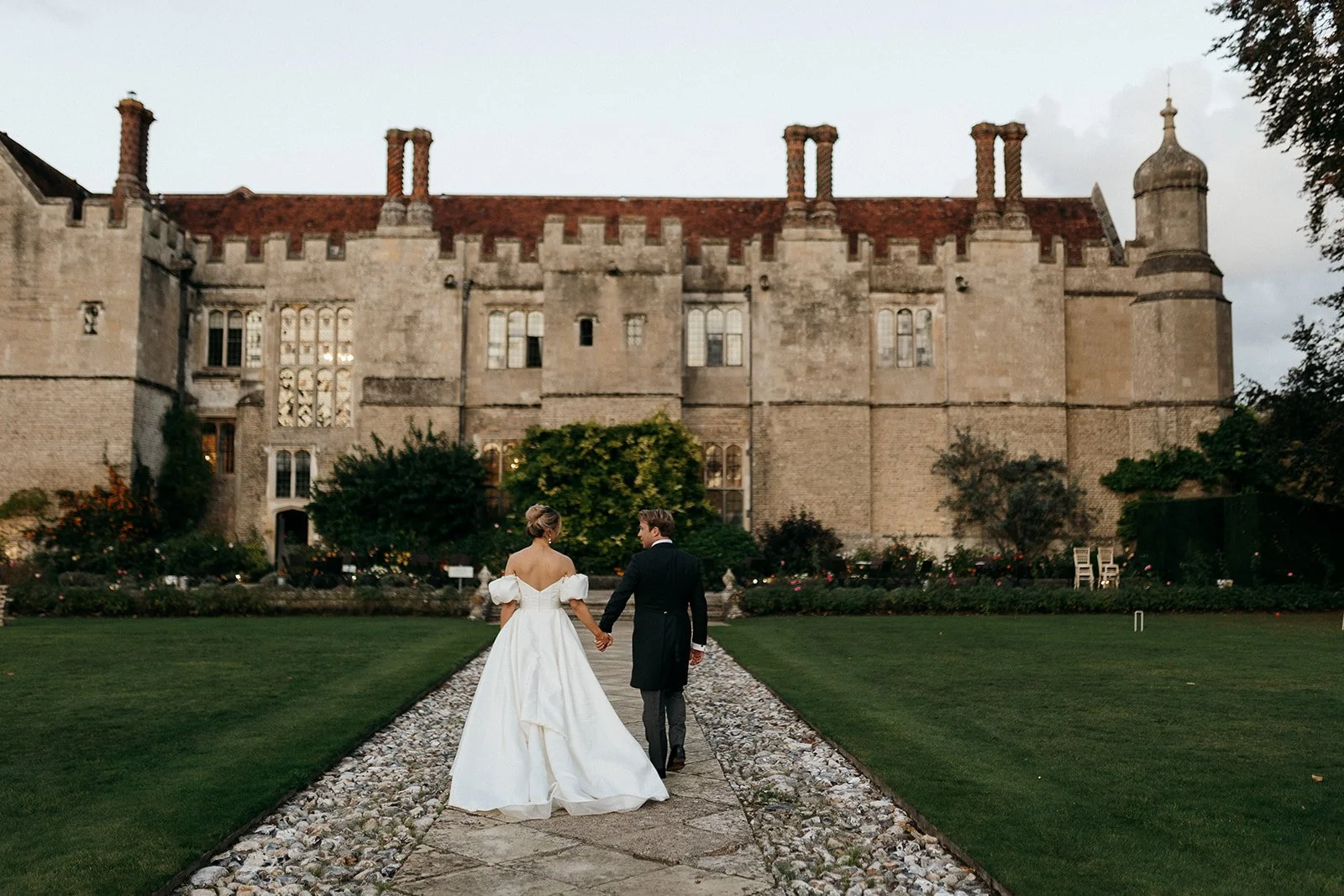 A bride and groom holding hands walking on a stone path towards a historic castle with a garden in the foreground during daytime.