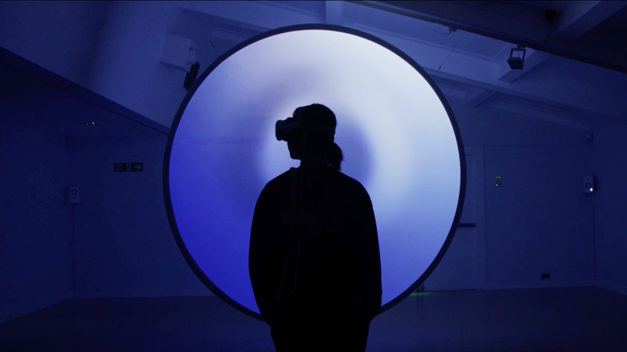 A silhouetted person wearing a VR headset stands facing a large glowing circular light installation in a dark gallery space, illuminated in shades of blue and white, creating a quiet, immersive atmosphere.