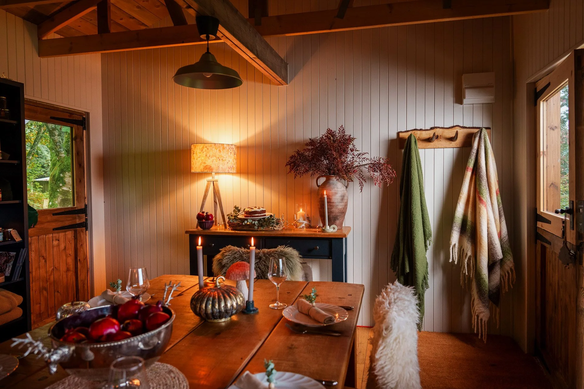 A cozy dining room with a wooden table set with candles, dishes, and a bowl of red apples. The room has wood-paneled walls, a sideboard with a table lamp, candles, and a large vase with a plant. Two open windows show green foliage outside.