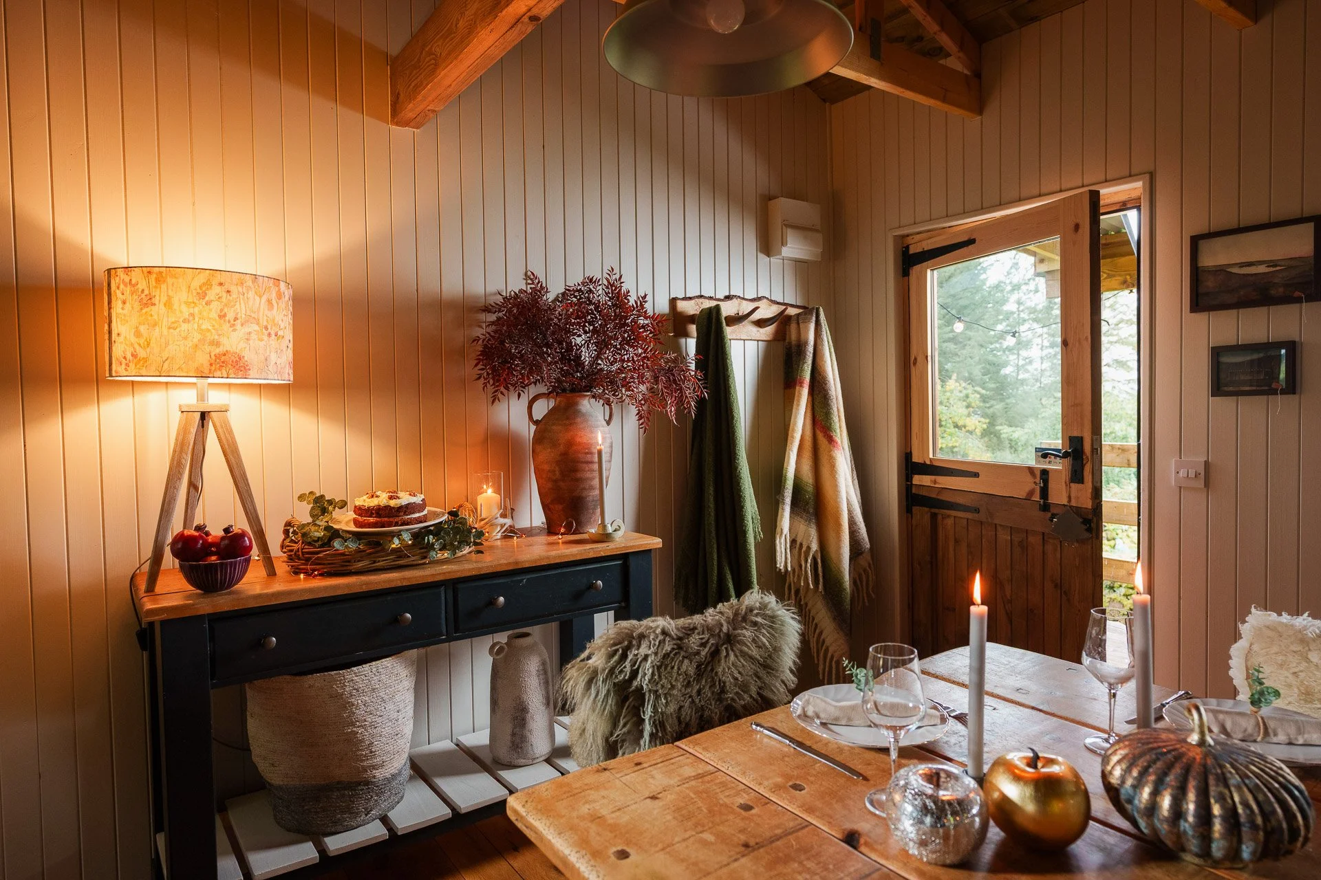 The Pantry set with candles, glasses, a pumpkin, and a gold apple. A sideboard decorated with a vase of red leaves, a cake, candles, and apples stands against the wall. The room has wood-panelled walls, a large window, and warm lighting.