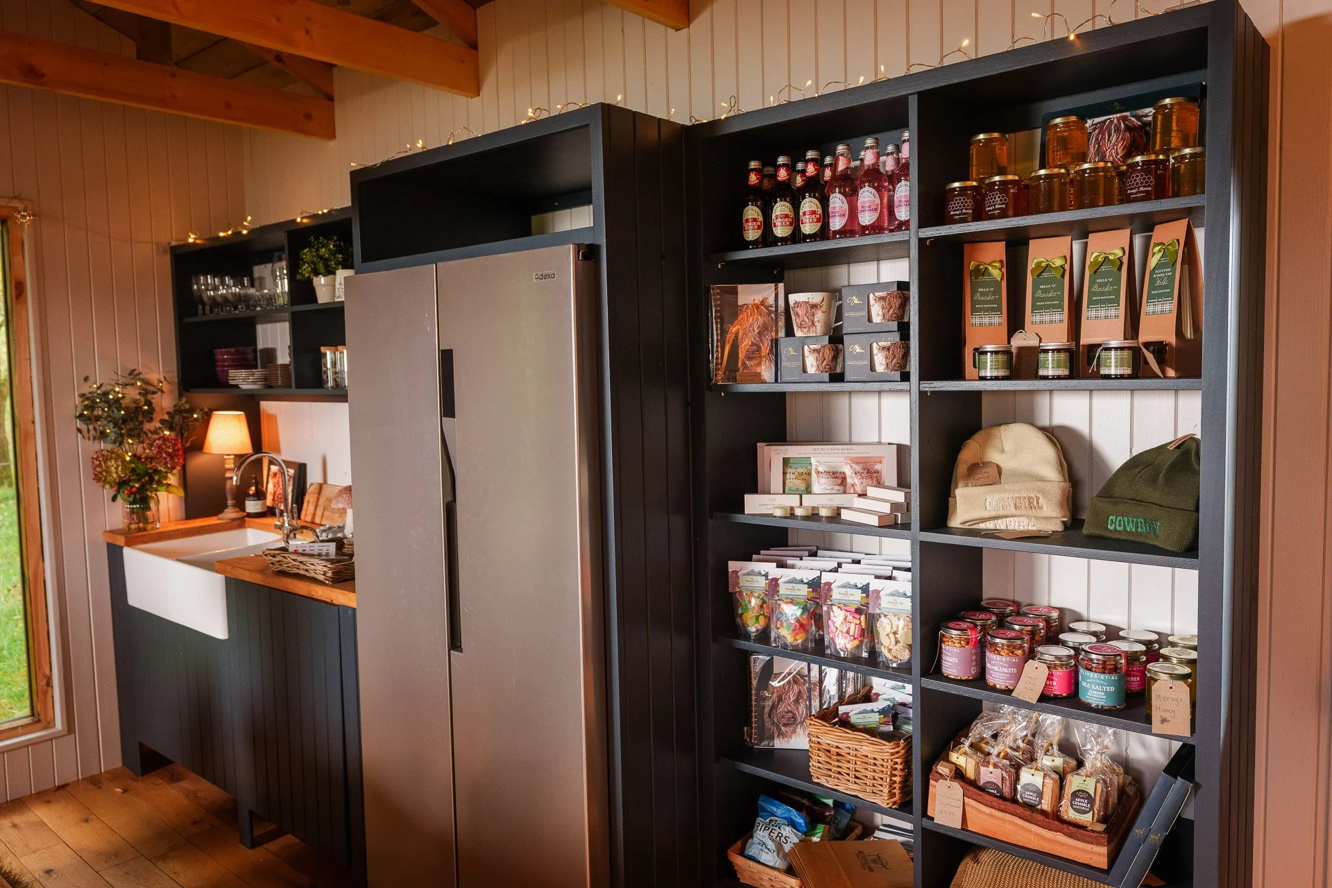 Interior view of a rustic shop with black shelving stocked with jars, boxes, bags, hats, and gift items, with a kitchen sink and counter to the left.