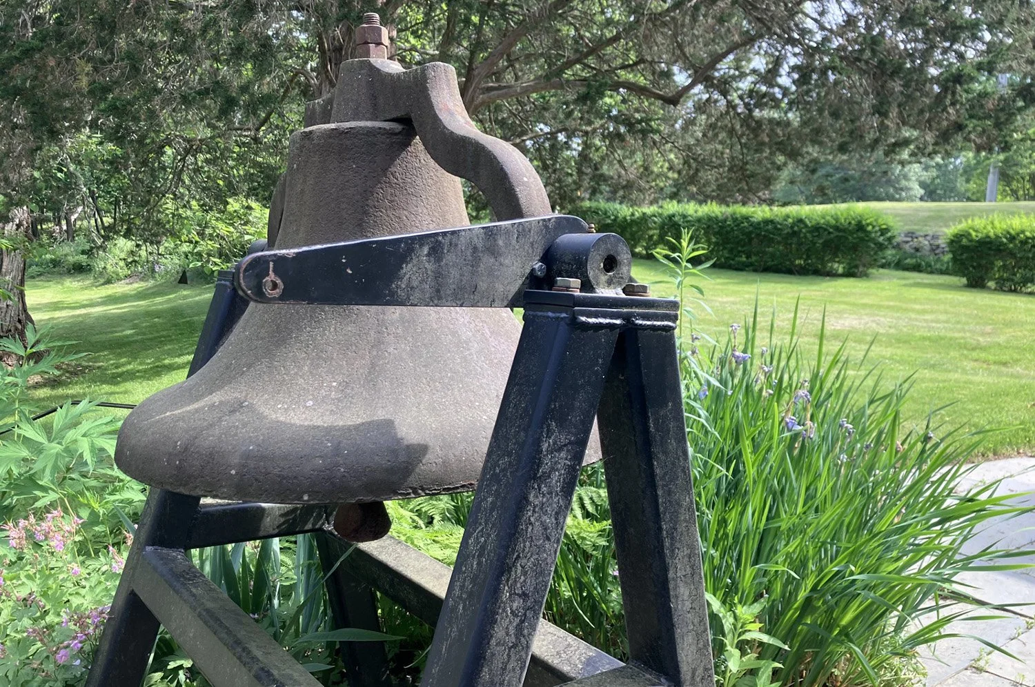 A weathered cast-iron bell suspended in a black steel frame in a monastery garden, with sunlit lawn, irises, ferns, and evergreen trees behind it.