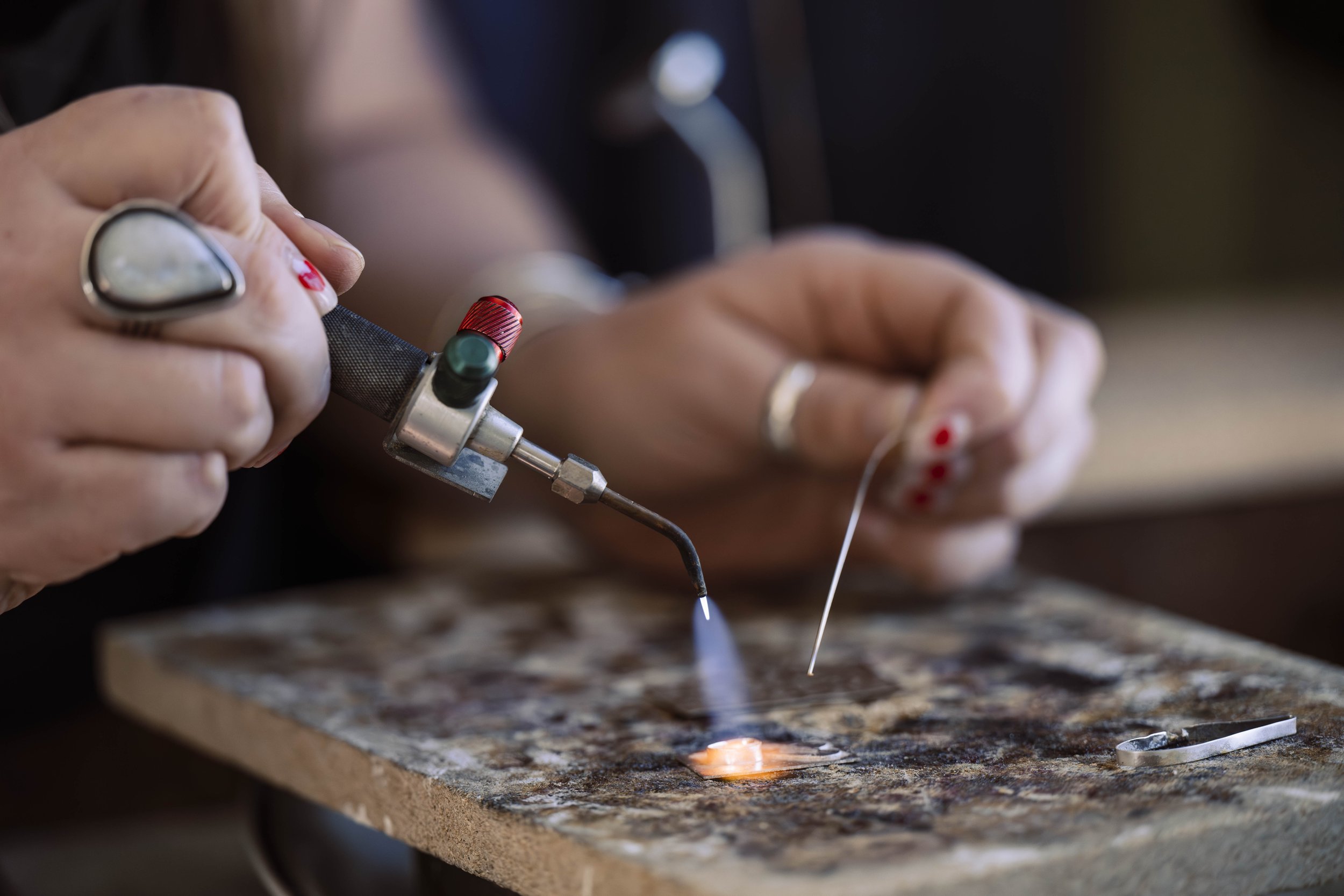 Person welding a small metal object using a torch and fine needle on a workbench.