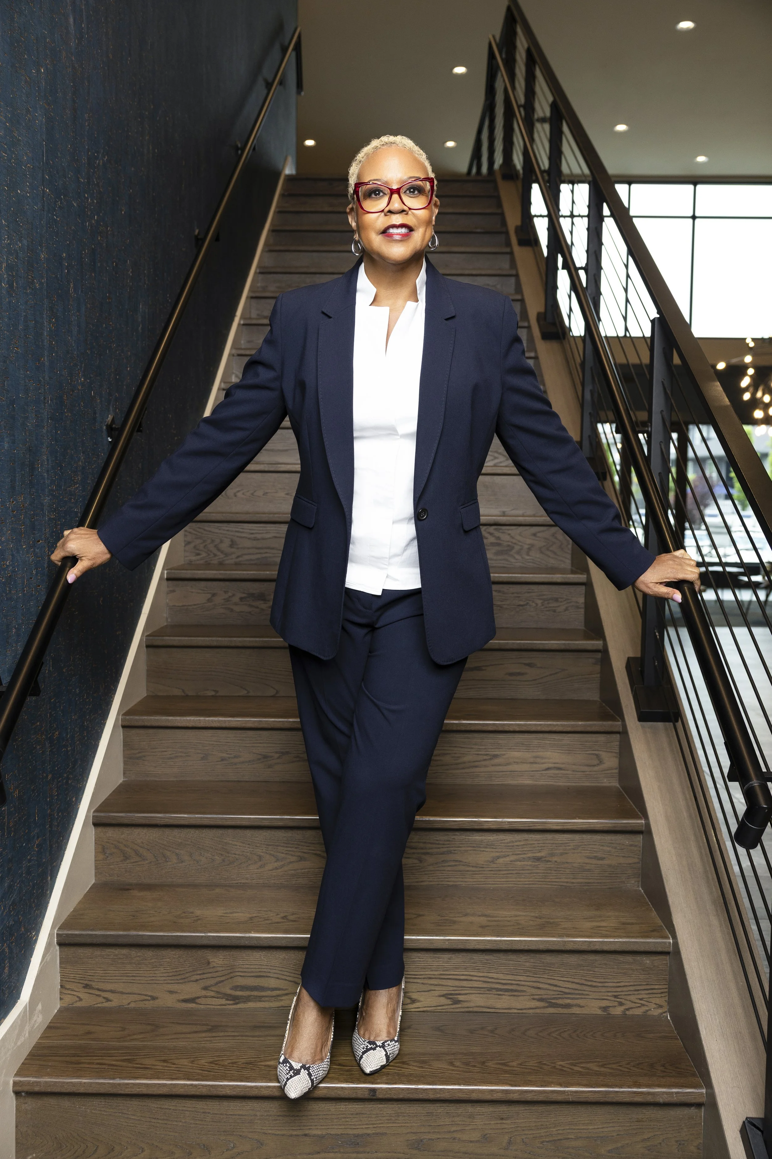 A confident middle-aged woman dressed in a navy suit with a white blouse, wearing red glasses, standing on a wooden staircase indoors, with a modern decor and large windows in the background.