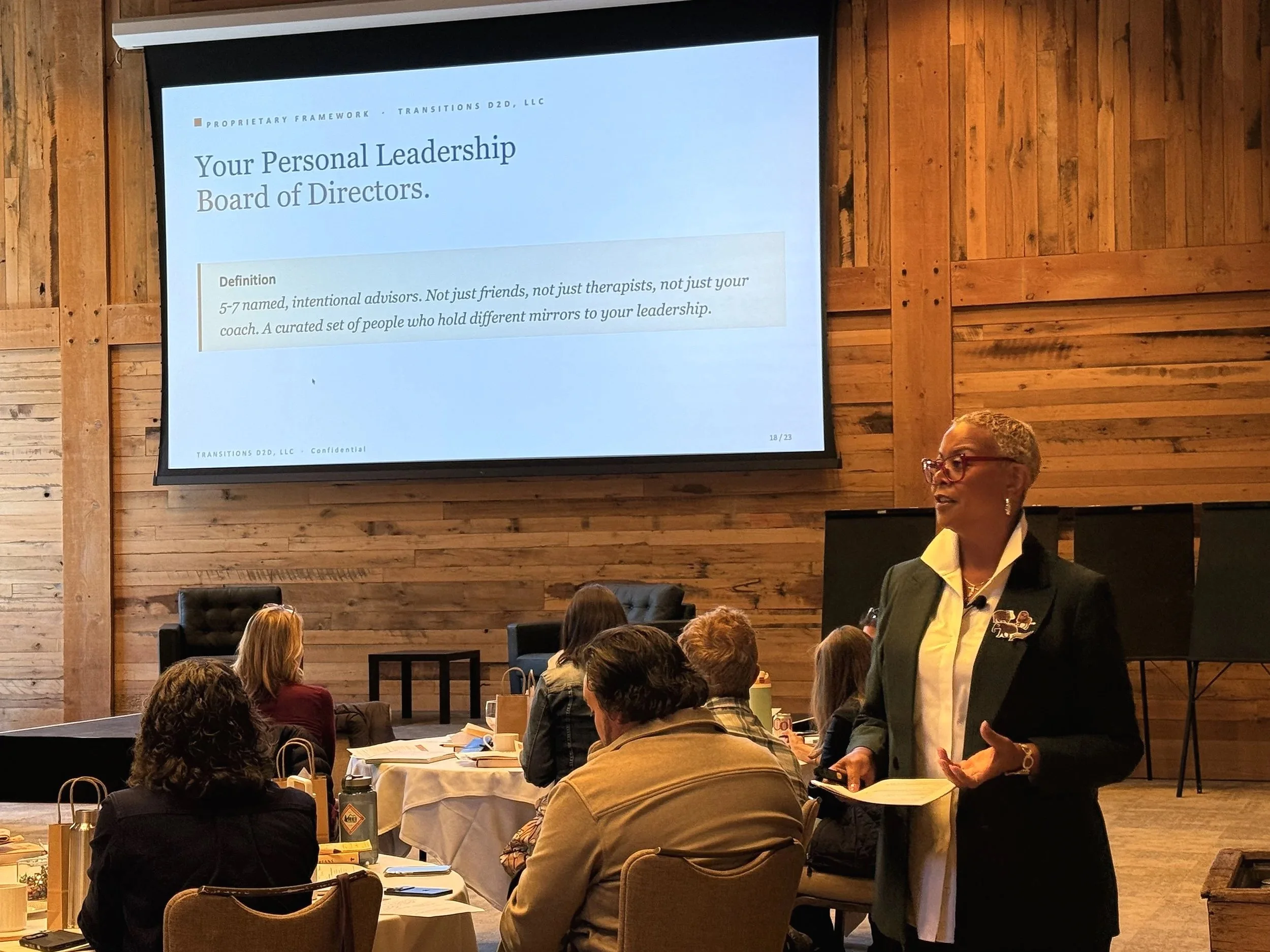 A woman in a blazer and glasses speaking during a presentation in a room with wooden walls. A large screen displays a slide titled 'Your Personal Leadership Board of Directors,' with text about advisors and mirrors for leadership.