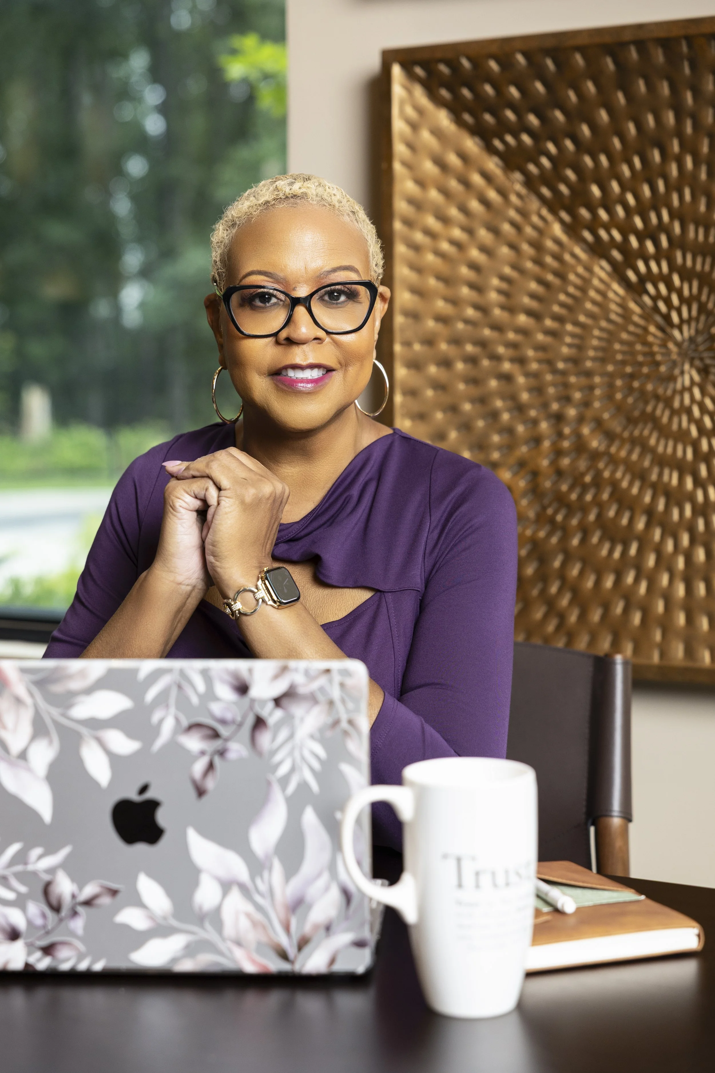 A woman with short blonde hair, glasses, and hoop earrings sitting at a table with a laptop, a white mug, and a notebook, in a cozy indoor setting with a wooden decorative panel behind her and greenery outside the window.