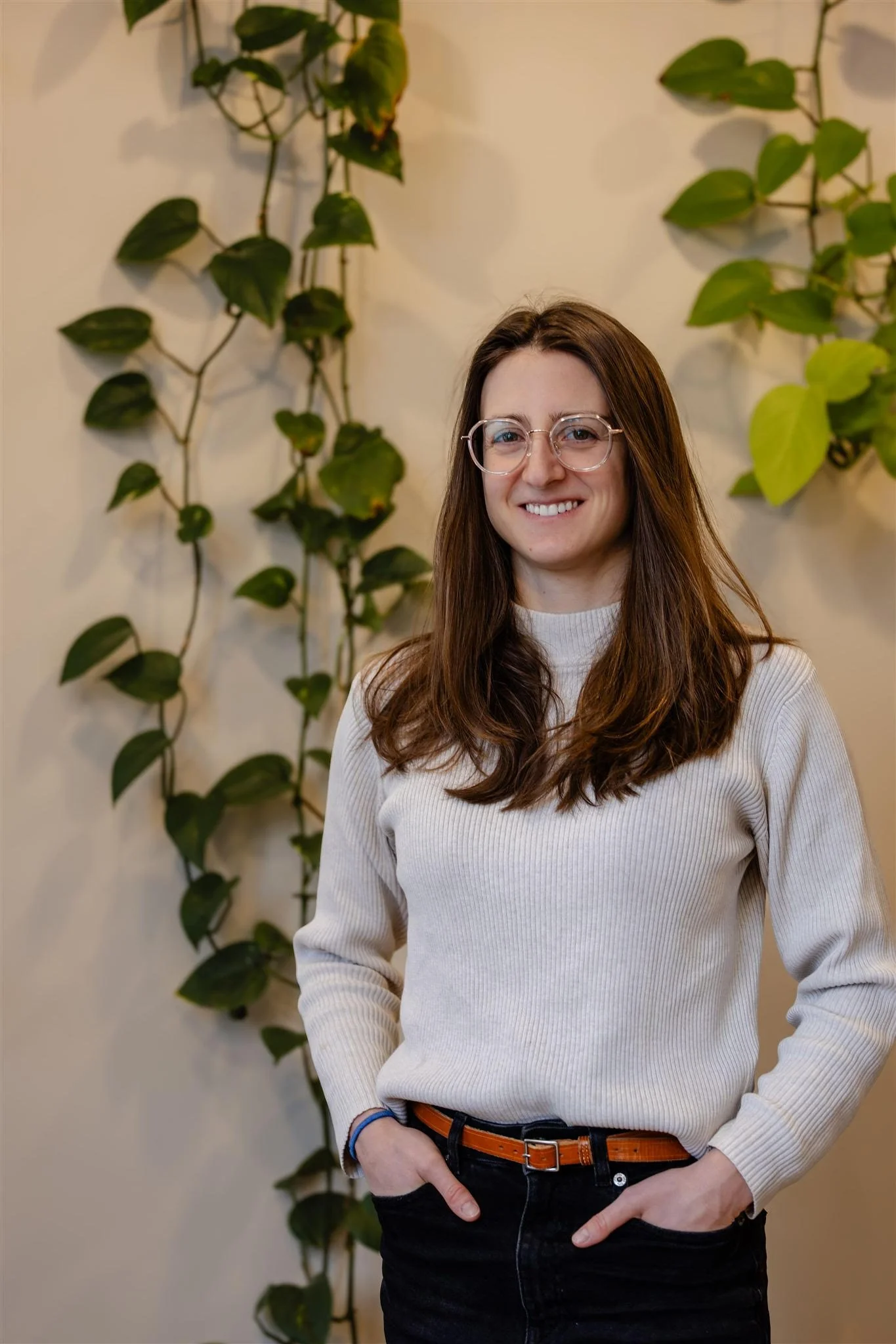 A woman with long brown hair wearing glasses, a cream-colored sweater, and black pants with a brown belt, standing in front of a wall with green leafy plants.