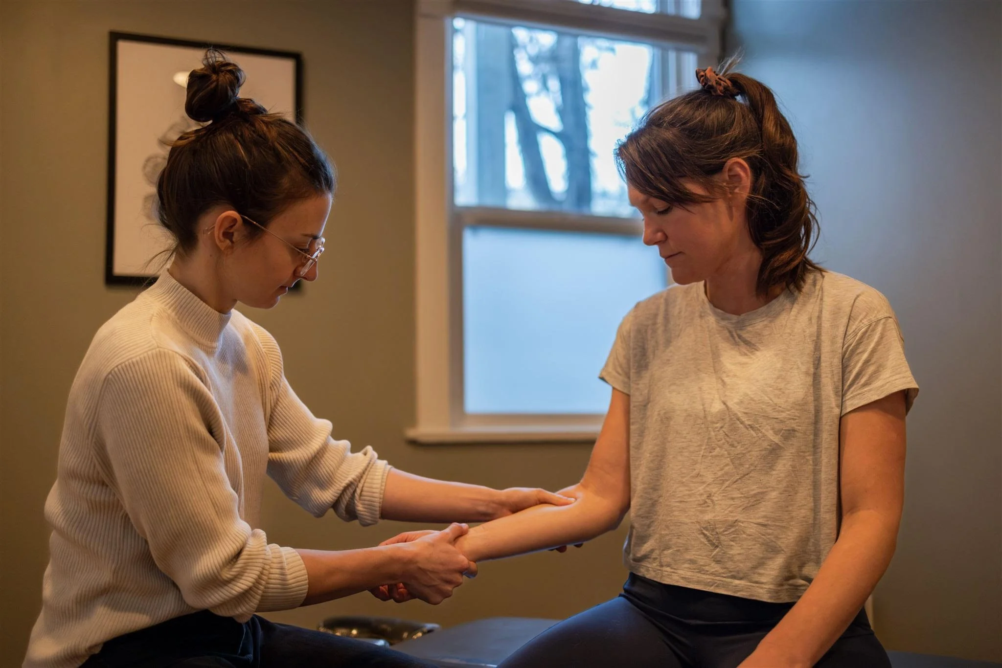 A woman assists another woman by holding her arm during a physical therapy session in a room with natural light.
