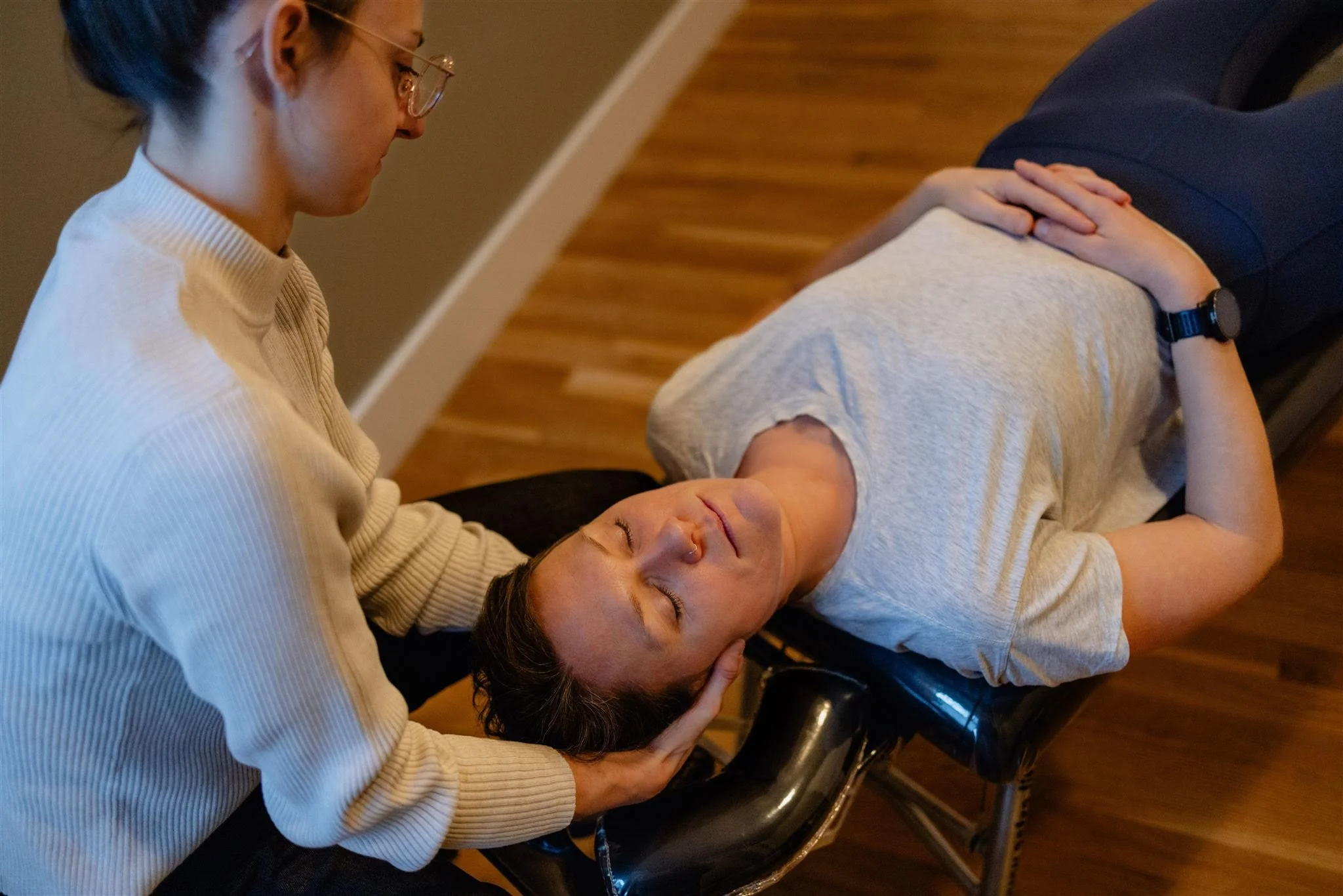 A woman lies on a chiropractic or massage table, receiving treatment or a check-up from a practitioner who is gently supporting her head with one hand.