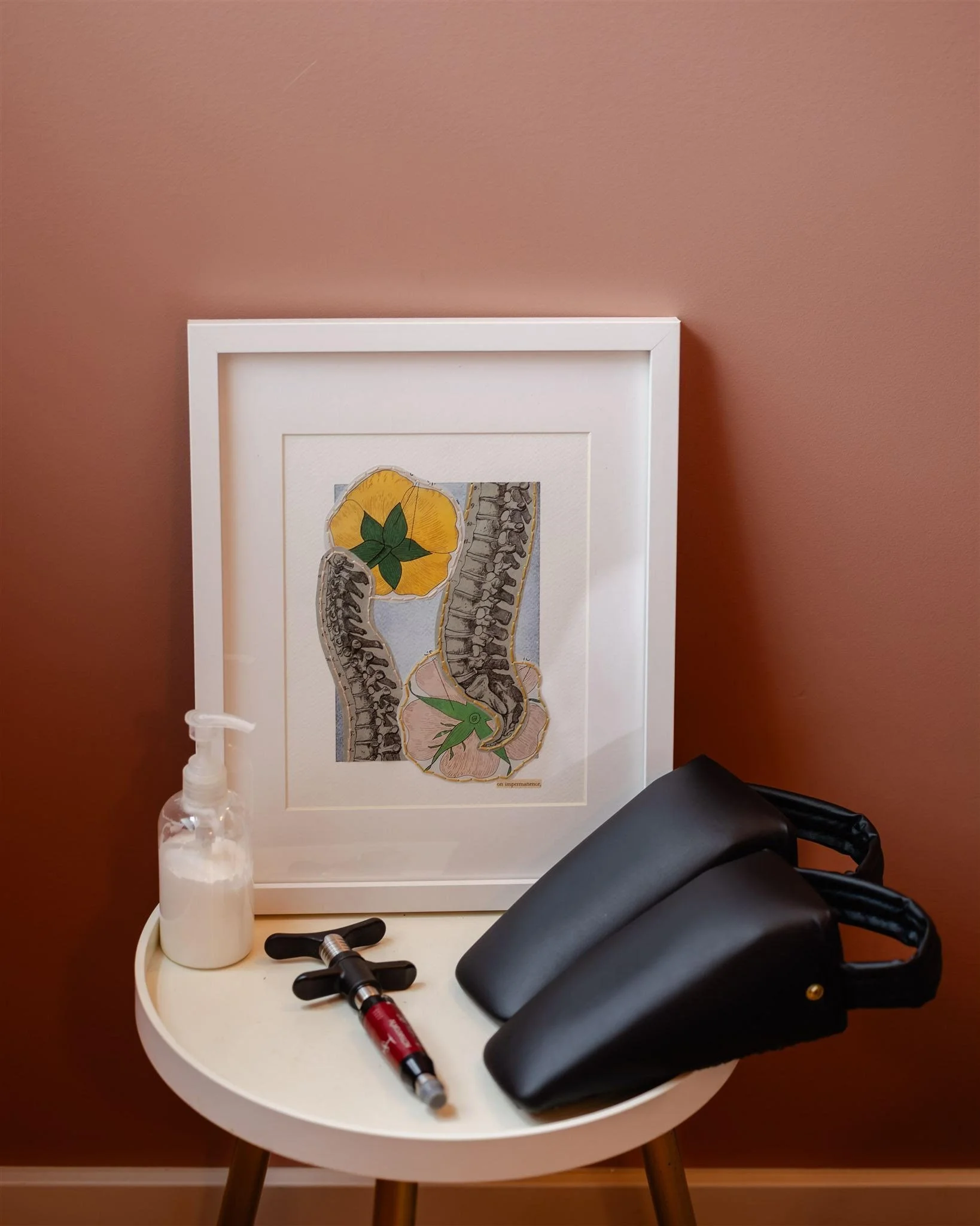 A small white table against a pink wall holds a framed illustration of a spine with yellow, pink, and gray elements, a white spray bottle, a black reflex hammer, and two black knee pads.