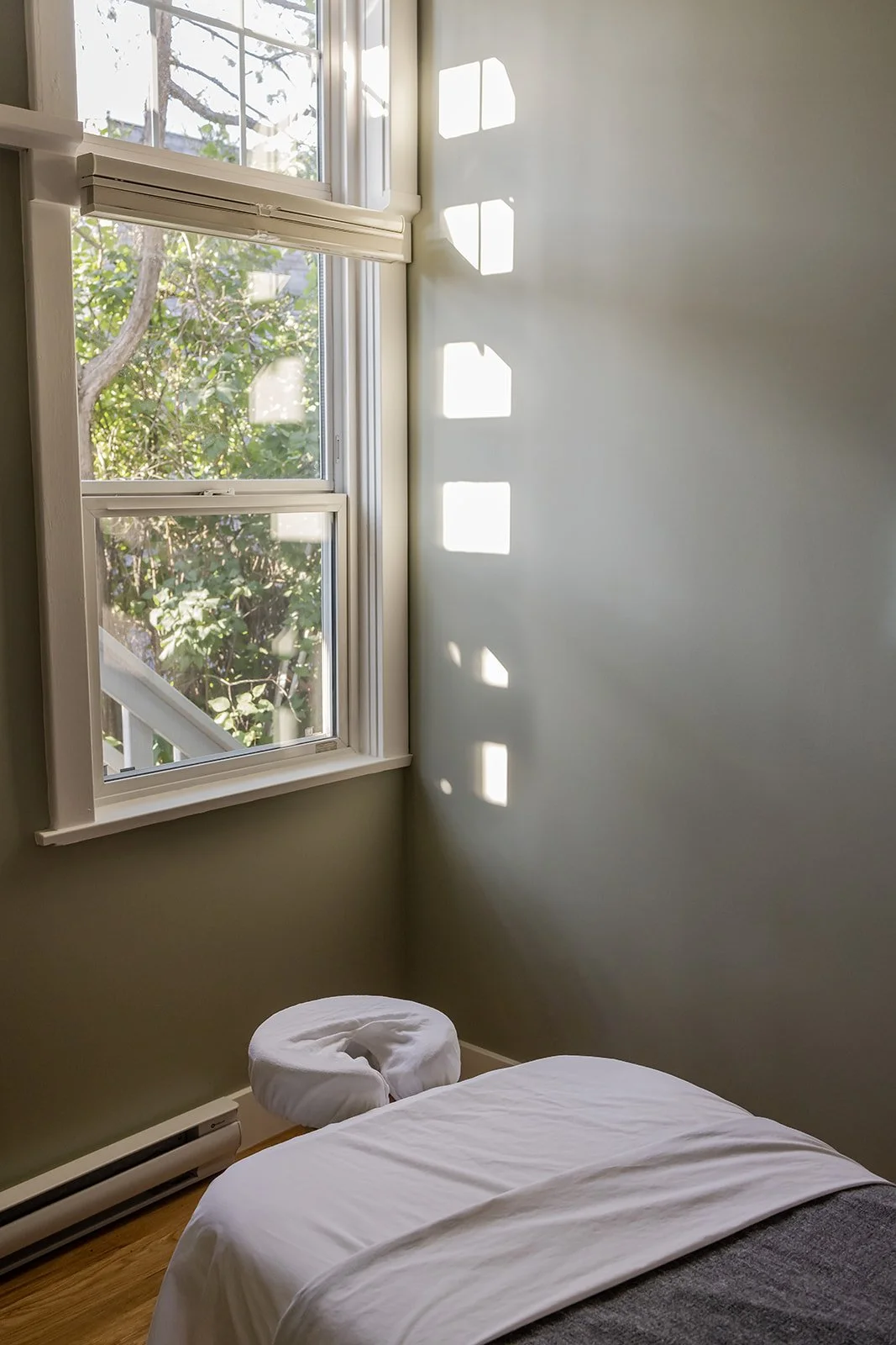 A bedroom with a window casting square-shaped shadows on the wall, a bed with white sheets, a folded towel or blanket on the bed, and a view of green foliage outside.