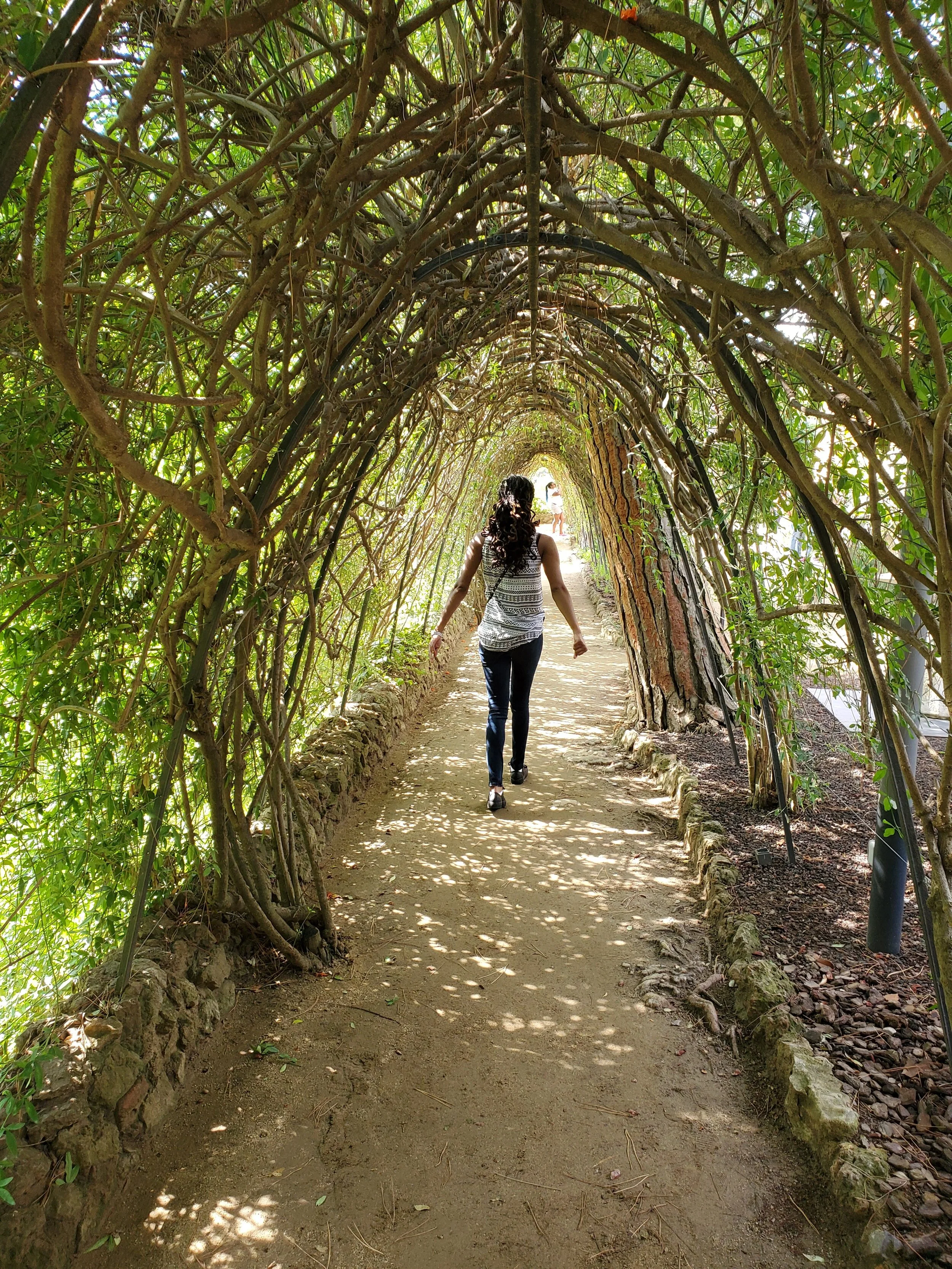 Black woman with long hair walking away through an archway of trees.