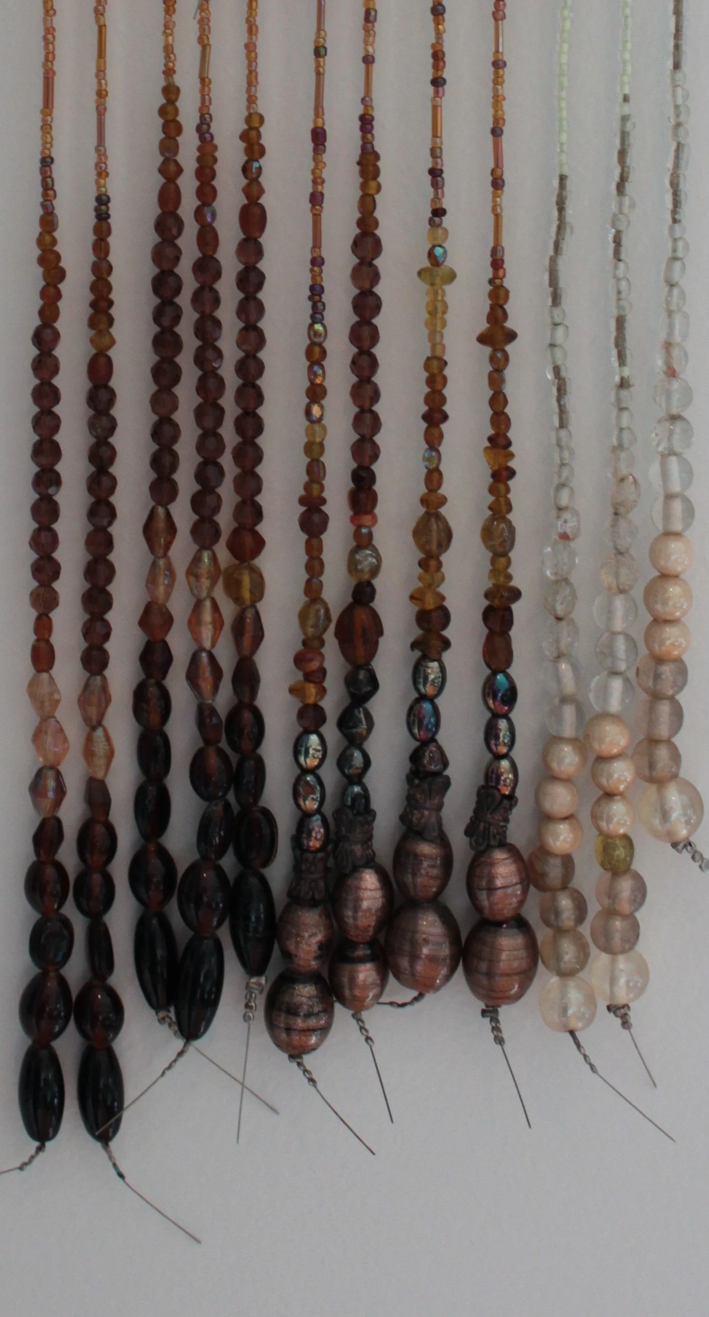 A display of multiple strands of beaded necklaces hanging against a white wall. The necklaces are made of various beads, ranging from dark brown to white, with some featuring iridescent or patterned beads.