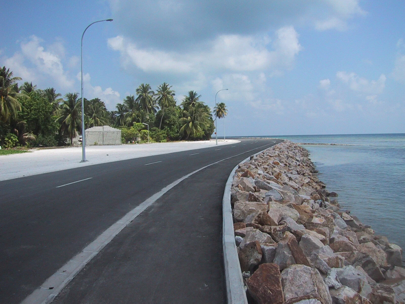 A paved coastal road runs alongside the ocean, with a rocky shoreline on one side and a line of palm trees on the other. The sky is partly cloudy with a mix of dark and light clouds.