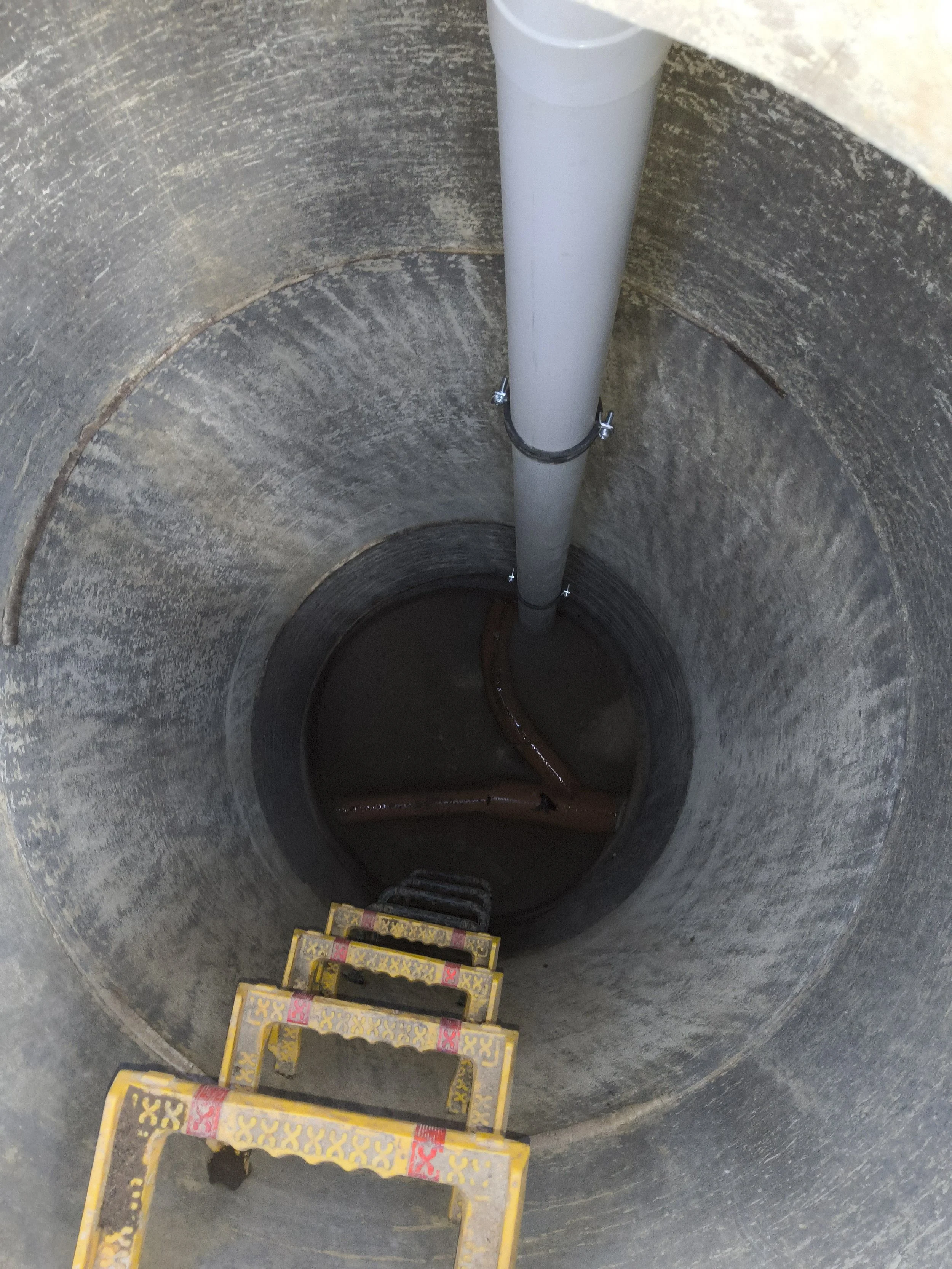 Inside a large, concrete well with a white pipe secured by metal clamps, and a yellow ladder at the bottom.