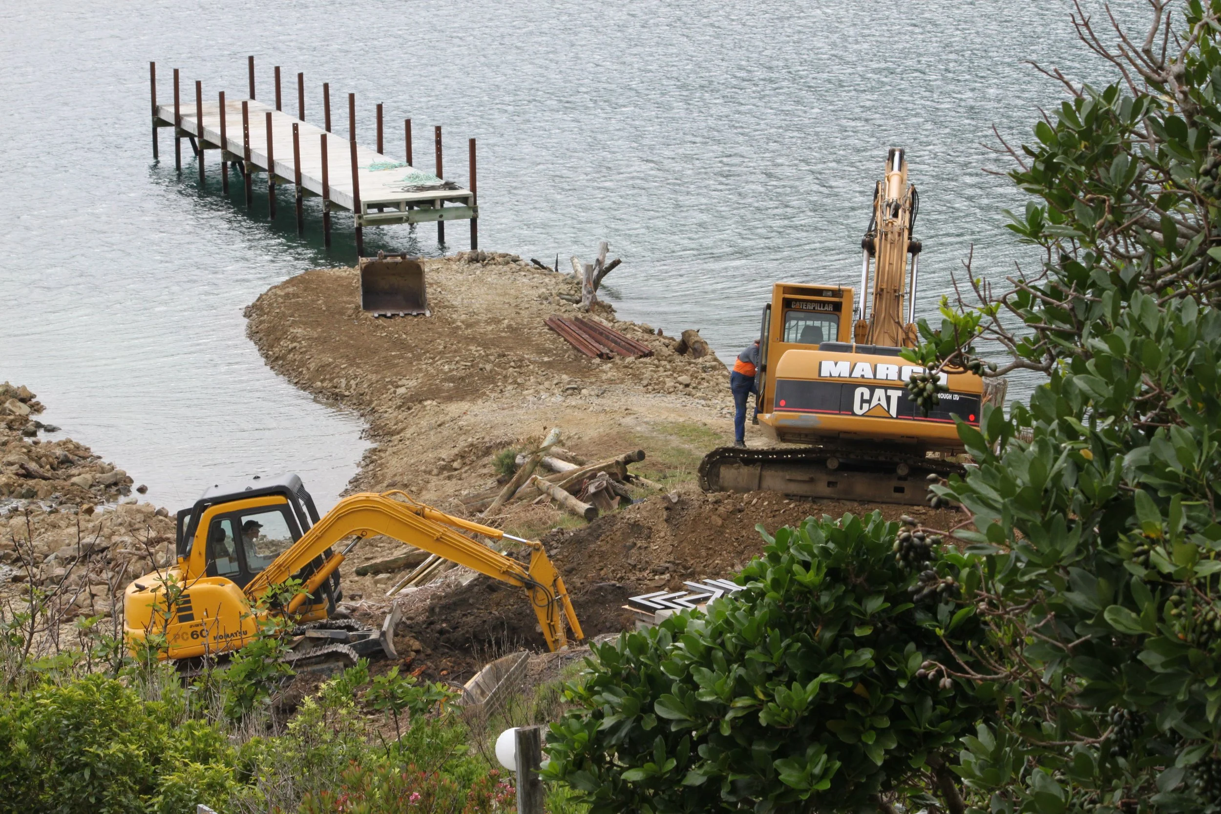 Construction site on the water with excavators working on land and a dock extending into the water, surrounded by green foliage.
