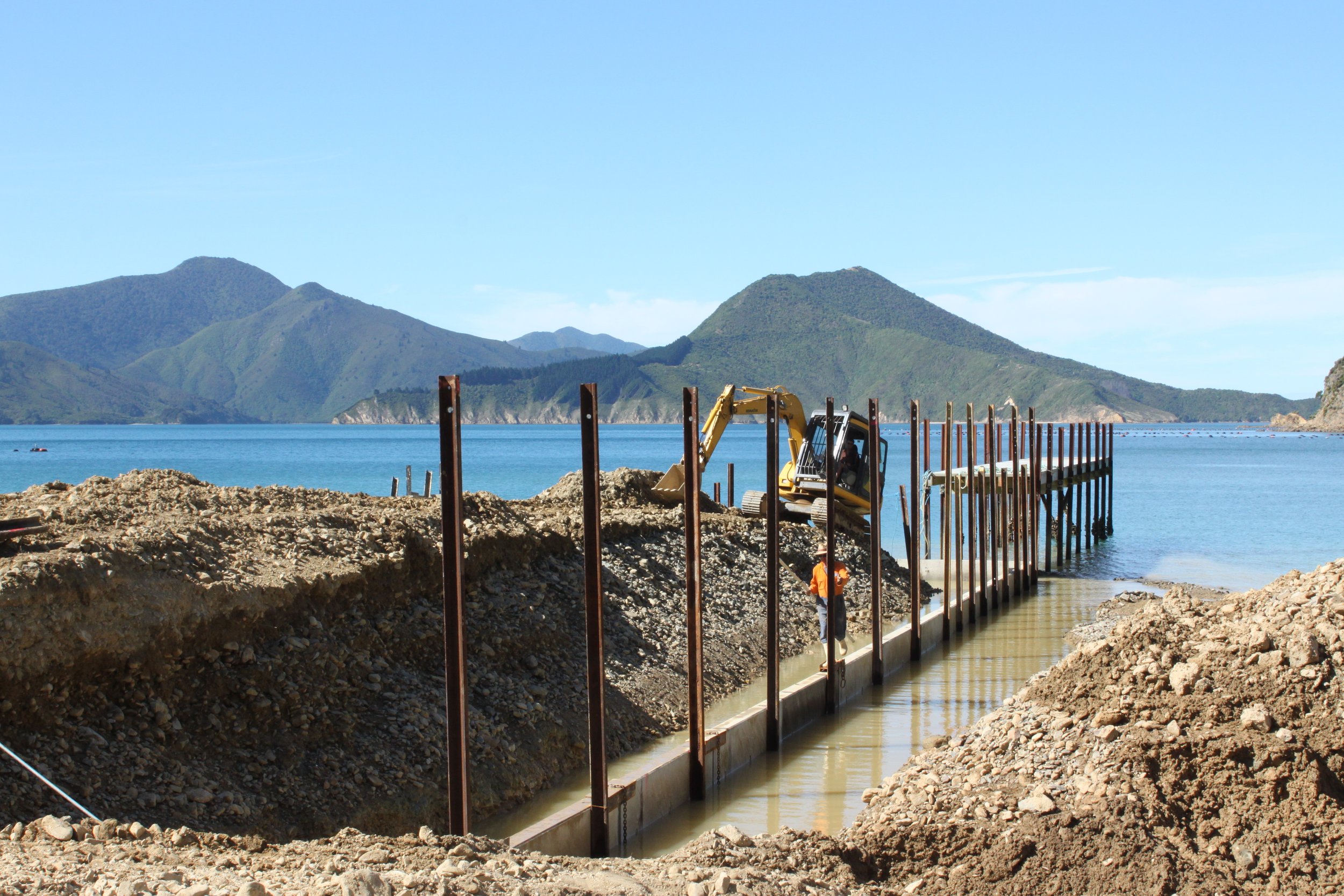 Construction site on a lakeshore with mountains in the background, a dirt trench, a yellow excavator, and metal pilings for a dock or pier.