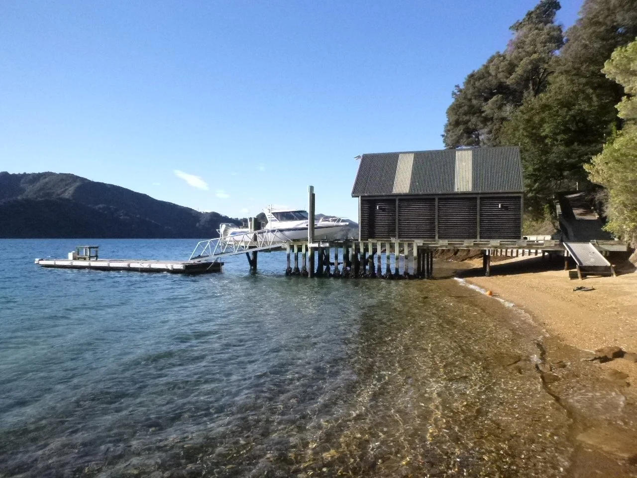 A dock with a boat on a ramp extending over water, a small hut on stilts, and trees on the shoreline with hills in the background under a clear blue sky.