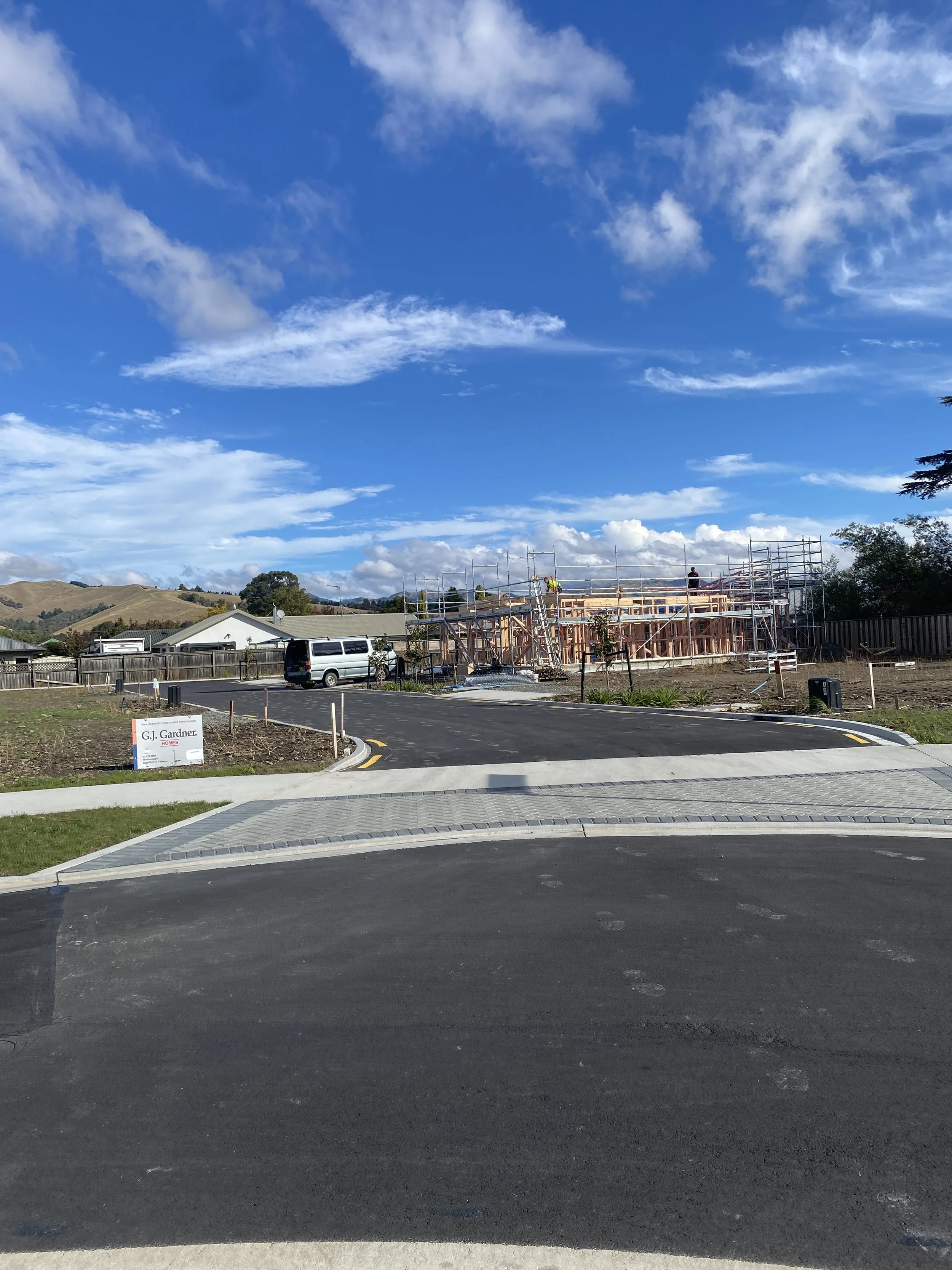 Construction site with building framework, scaffolding, workers, and a white van, under a partly cloudy blue sky with rolling hills in the background.