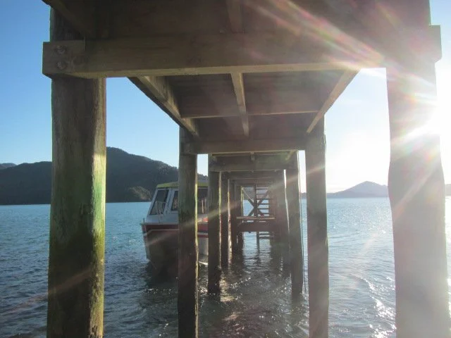 View underneath a wooden dock over water with a boat docked to the side and mountains in the background, sunlight glare in the upper right corner.