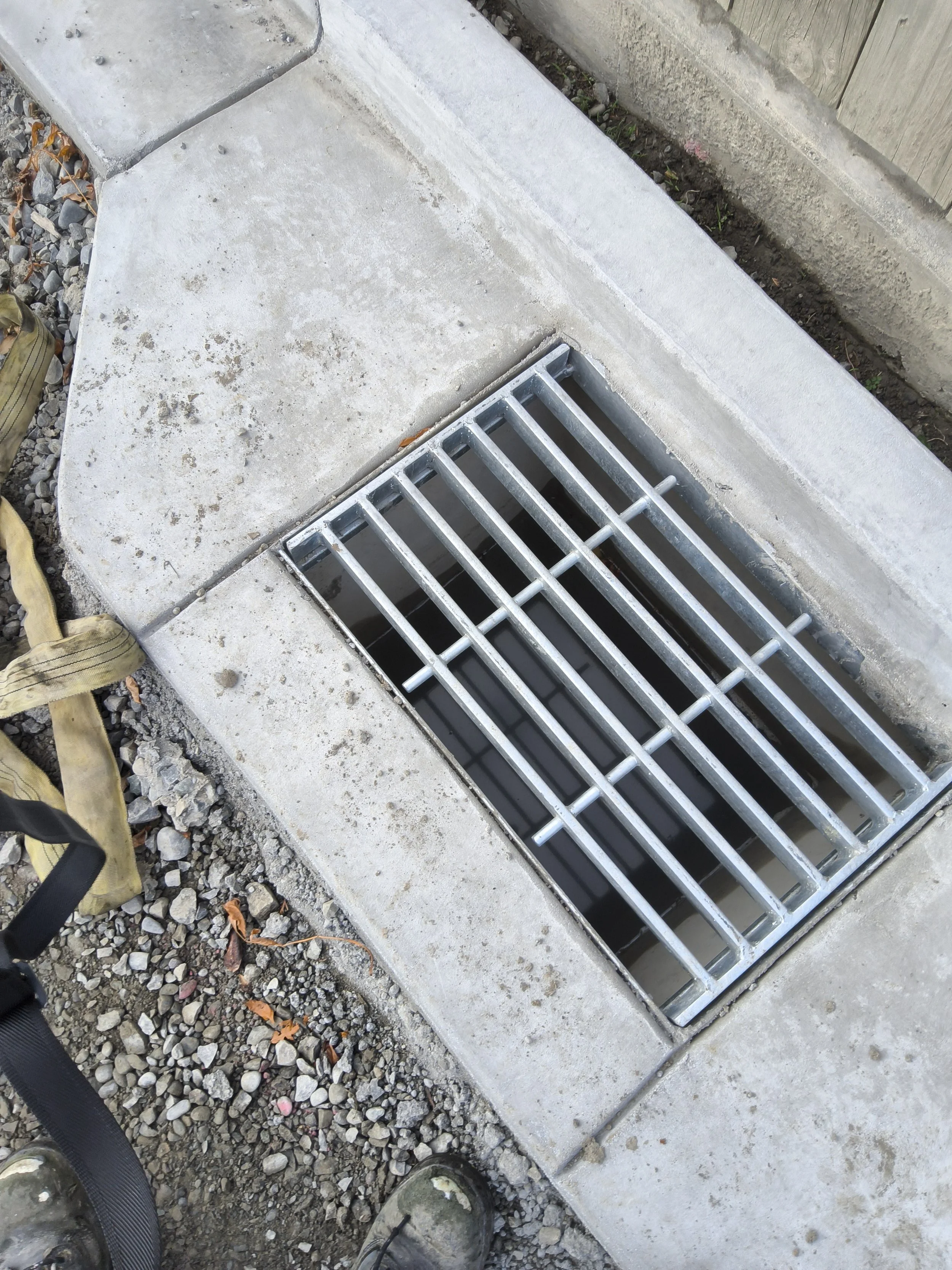 Close-up of a drainage grate installed in concrete sidewalk with gravel on the side and a wooden fence in the background.