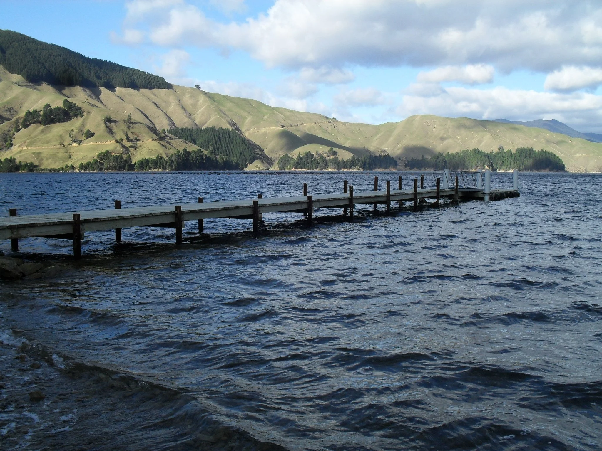 A lake with a wooden dock extending into the water, surrounded by hills covered in green grass and trees, under a partly cloudy sky.