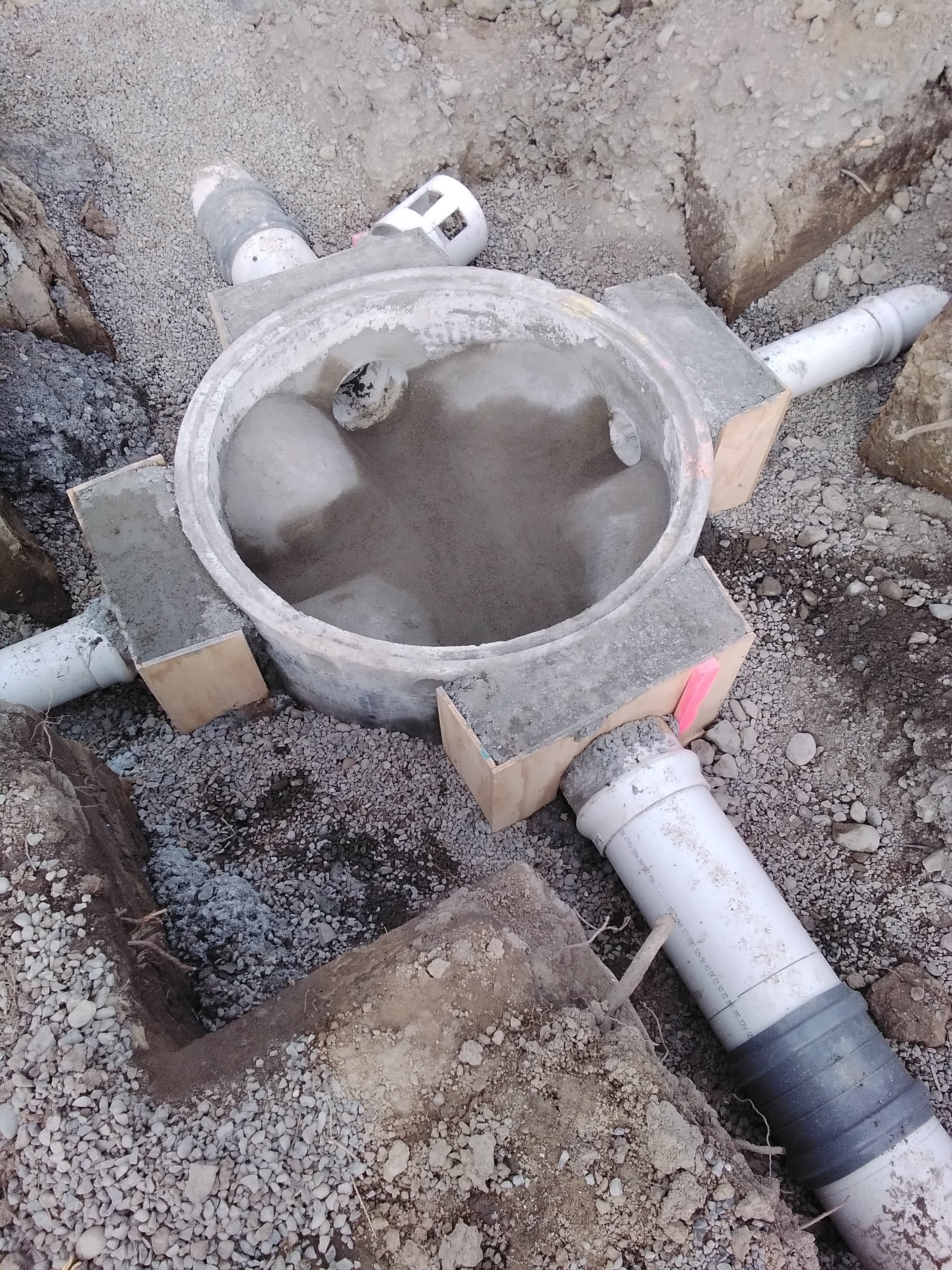 A construction site showing a pipe fitting with PVC pipes underground, surrounded by dirt and gravel.