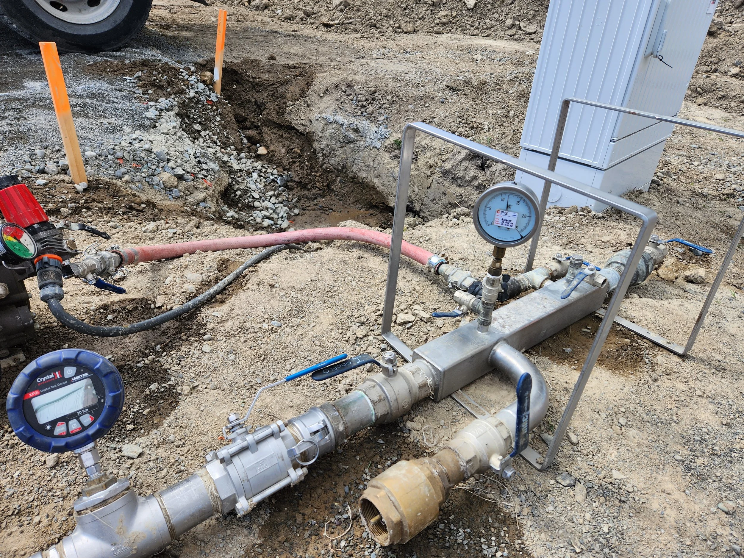 View of construction site with exposed pipes, a pressure gauge, and safety markers in dirt and gravel ground.
