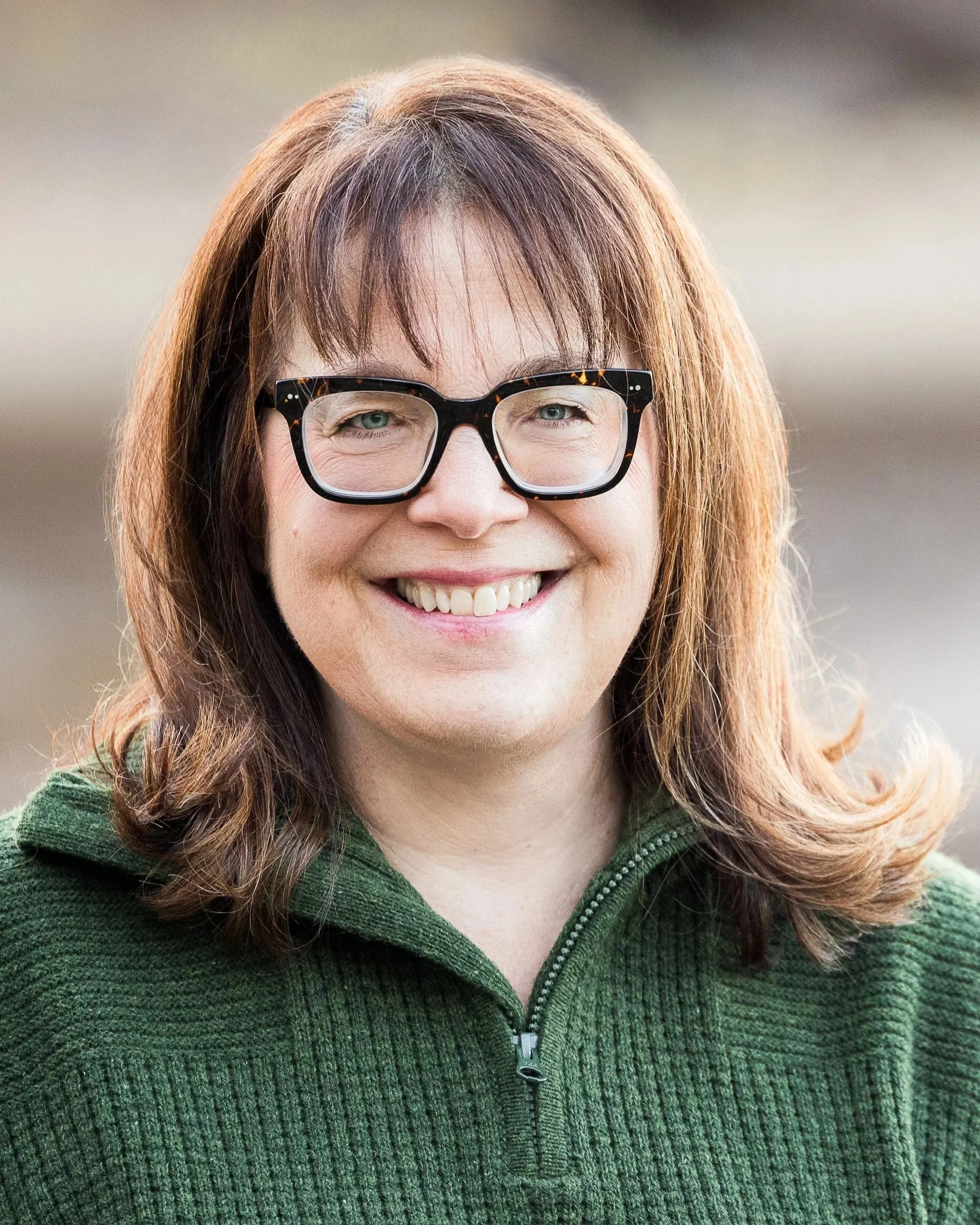 A woman with shoulder-length light brown hair, wearing glasses and a green zip-up sweater, smiling outdoors.