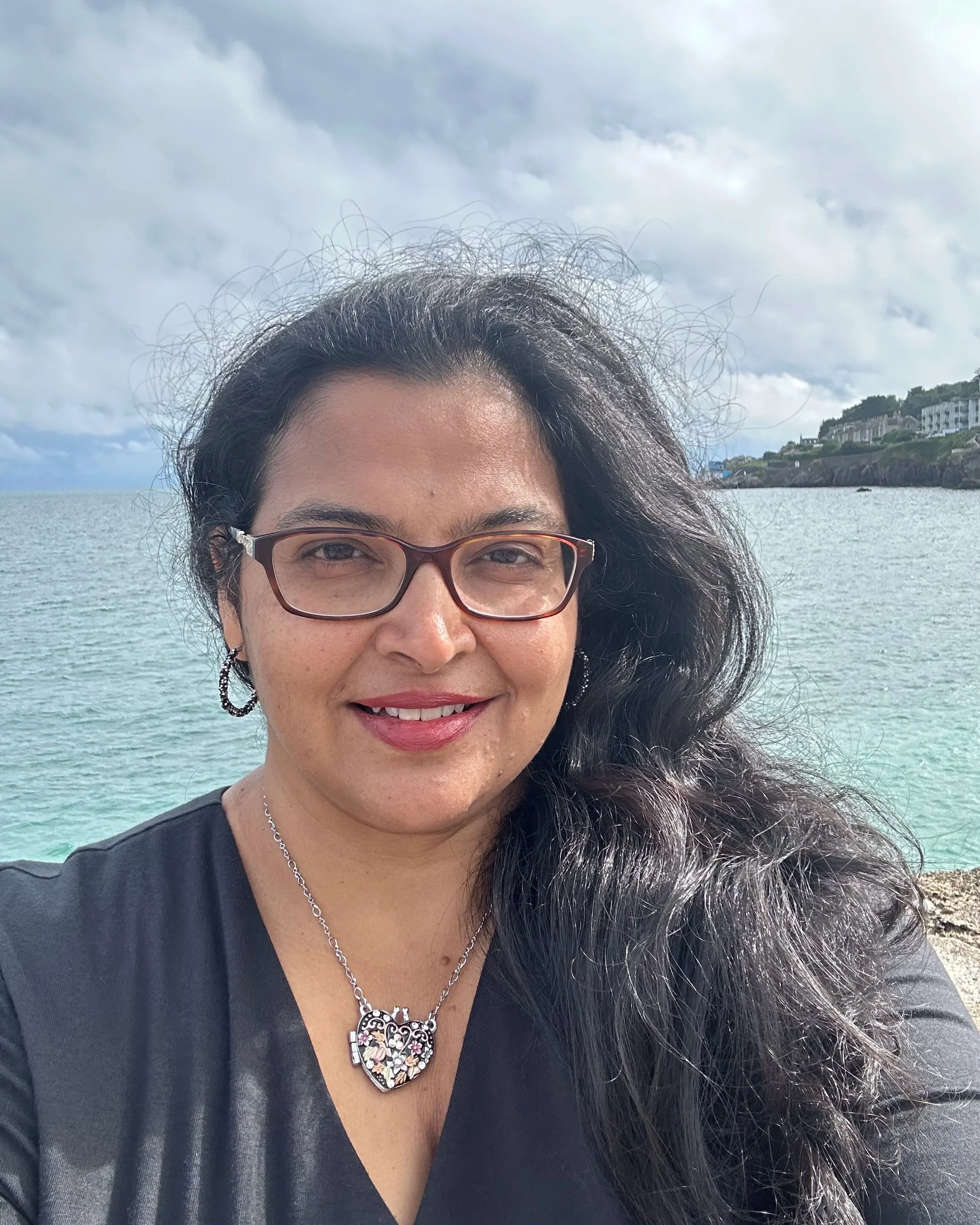 A woman with dark hair, glasses, and earrings smiling at the camera, standing by the ocean with waves, rocky shoreline, and houses on a hill in the background during the daytime with cloudy skies.