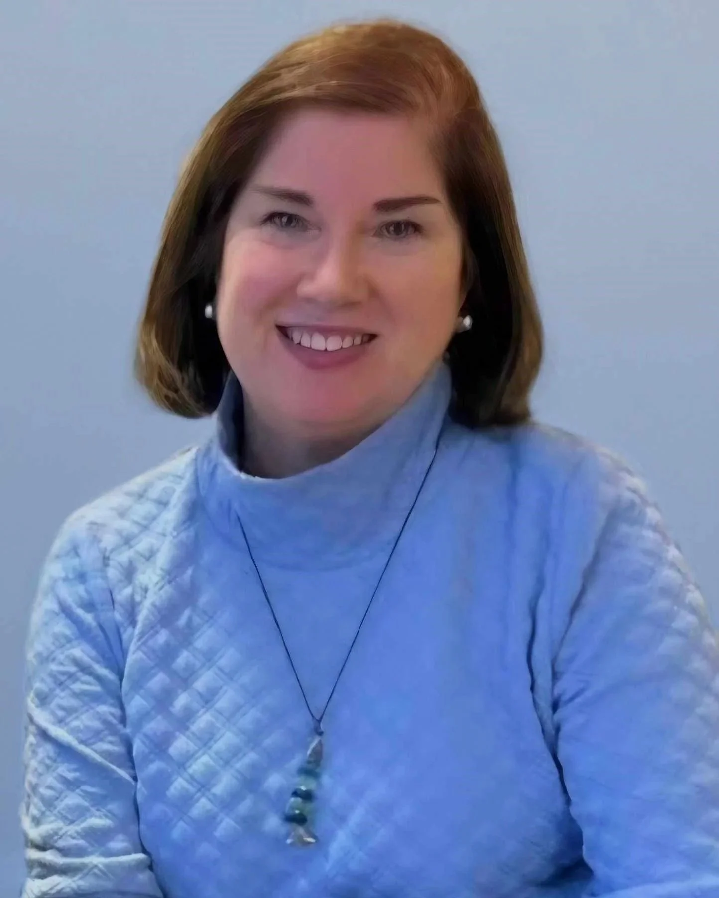 A woman with shoulder-length reddish-brown hair, smiling, wearing a light blue quilted turtleneck sweater, pearl earrings, and a necklace with a green and white pendant, against a plain blue-gray background.
