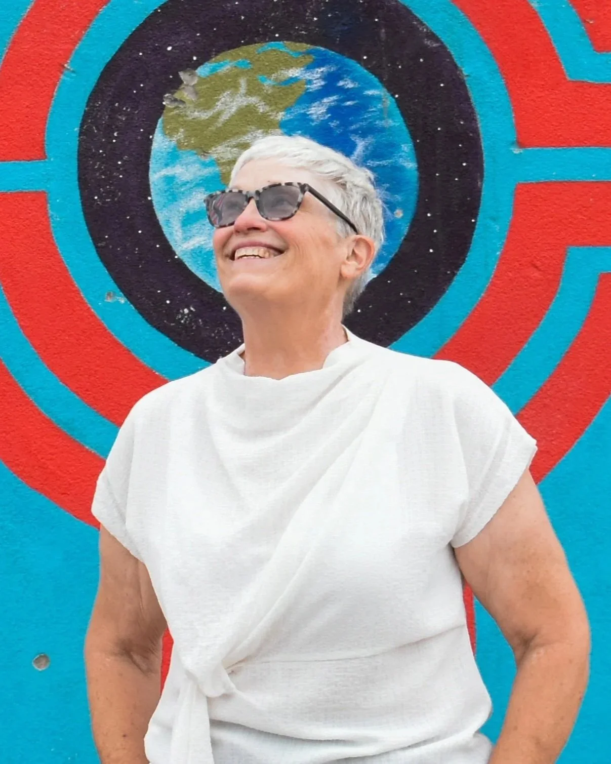 Smiling woman with short gray hair, wearing sunglasses and a white top, standing in front of a colorful mural featuring the Earth and a red and blue target design.