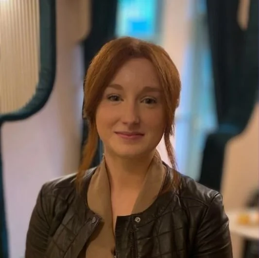 Woman in a red jacket standing next to a black and white dog in a wooded area with leaf-covered ground and leafless trees.