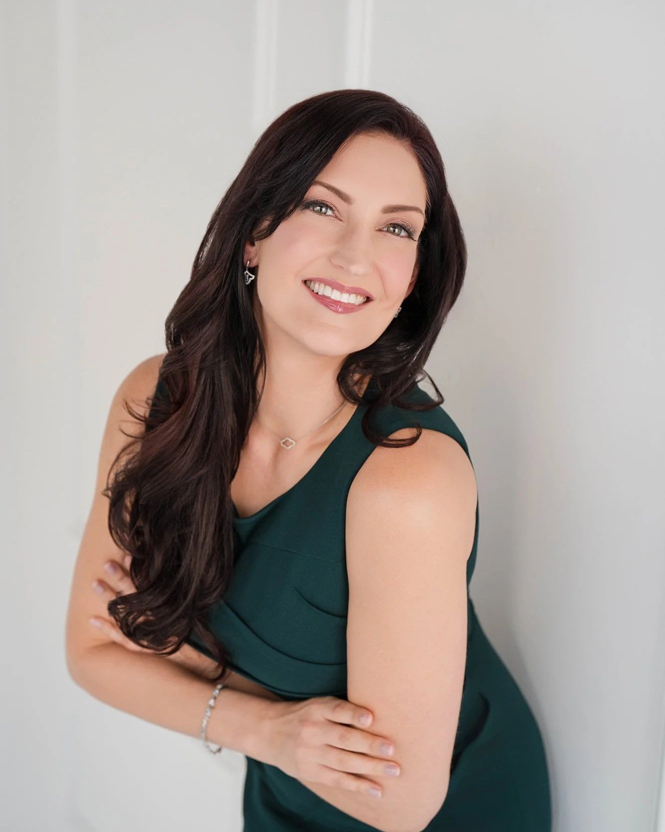 A woman with long dark hair and a bright smile, dressed in a dark green sleeveless dress, standing against a plain light-colored background.