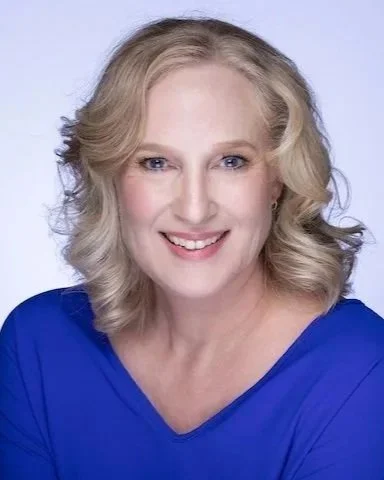 A woman with blonde, wavy hair styled in loose curls, smiling, wearing a blue top against a light background.
