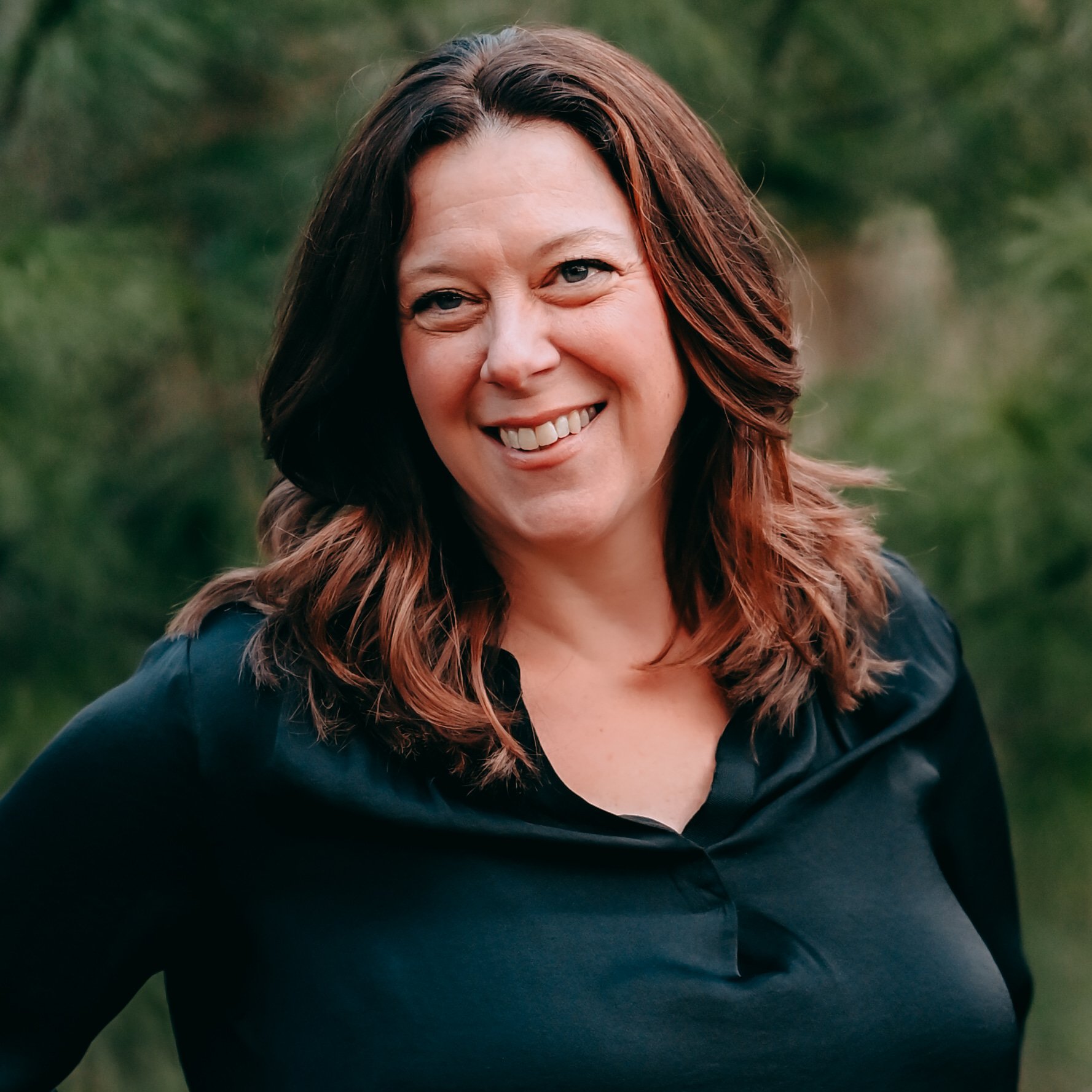 A smiling woman with long red hair wearing a black top and a gold necklace.