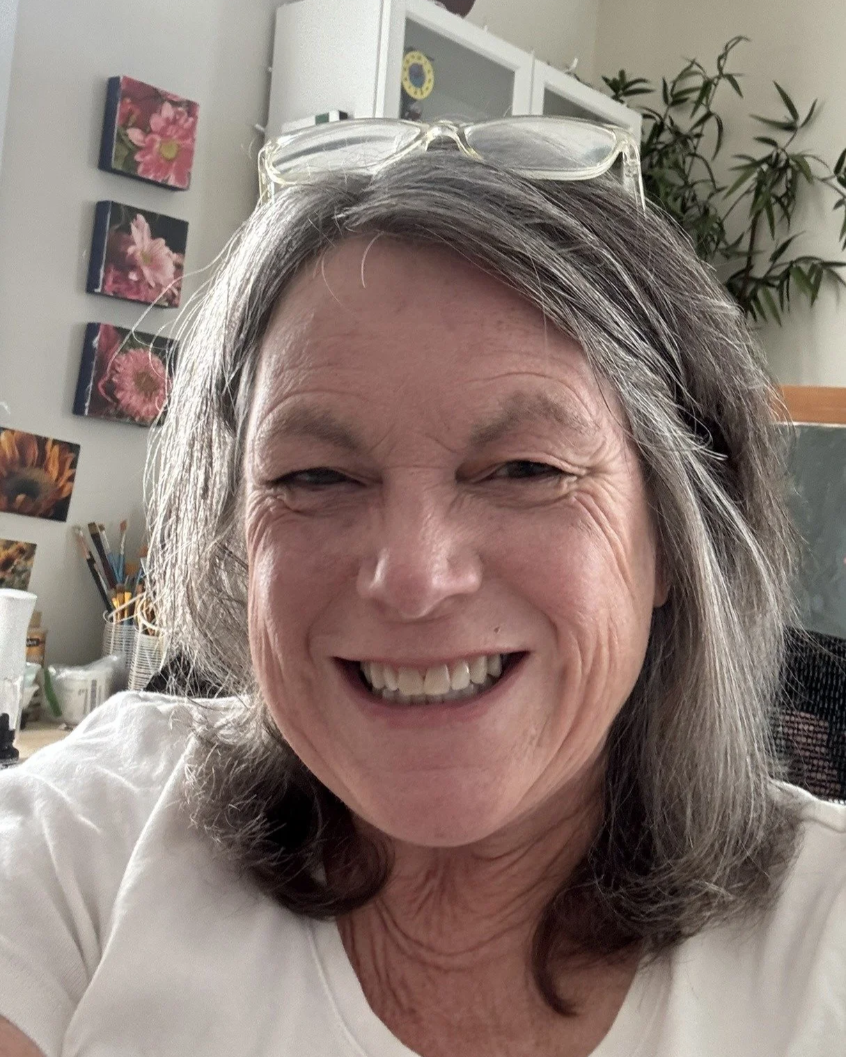 A smiling woman with glasses on top of her head taking a selfie indoors, with artwork and plants in the background.