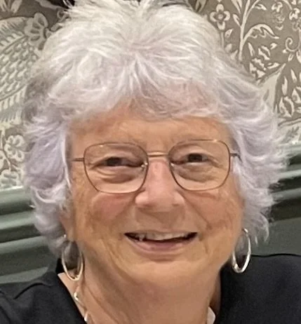 Smiling elderly woman with white hair, glasses, and hoop earrings, wearing a black top, in front of a patterned background.