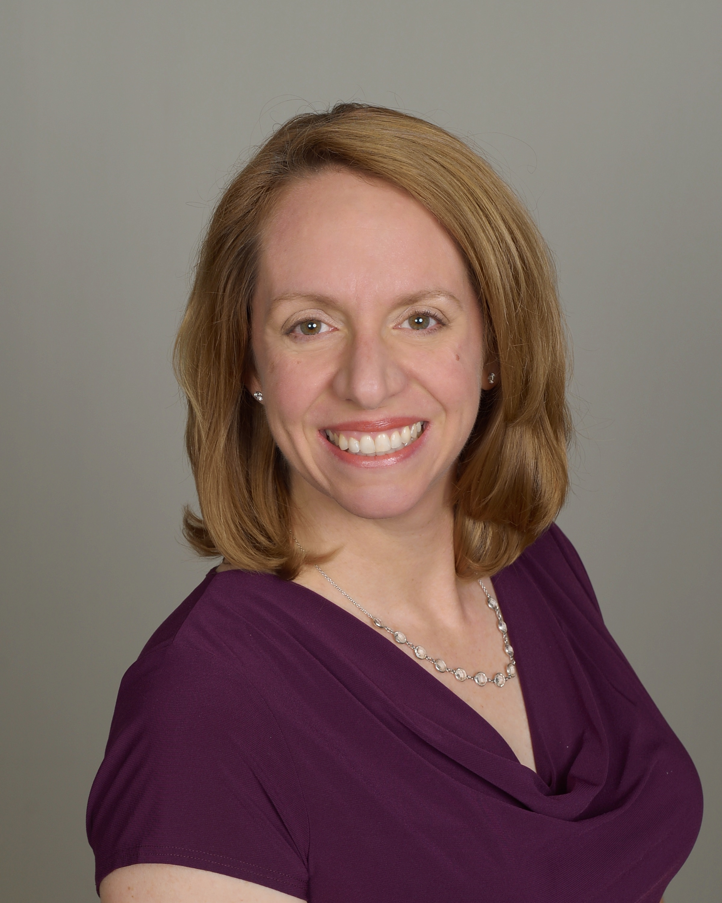Headshot of a smiling woman with shoulder-length red hair wearing a purple top and a necklace, posed against a plain background.