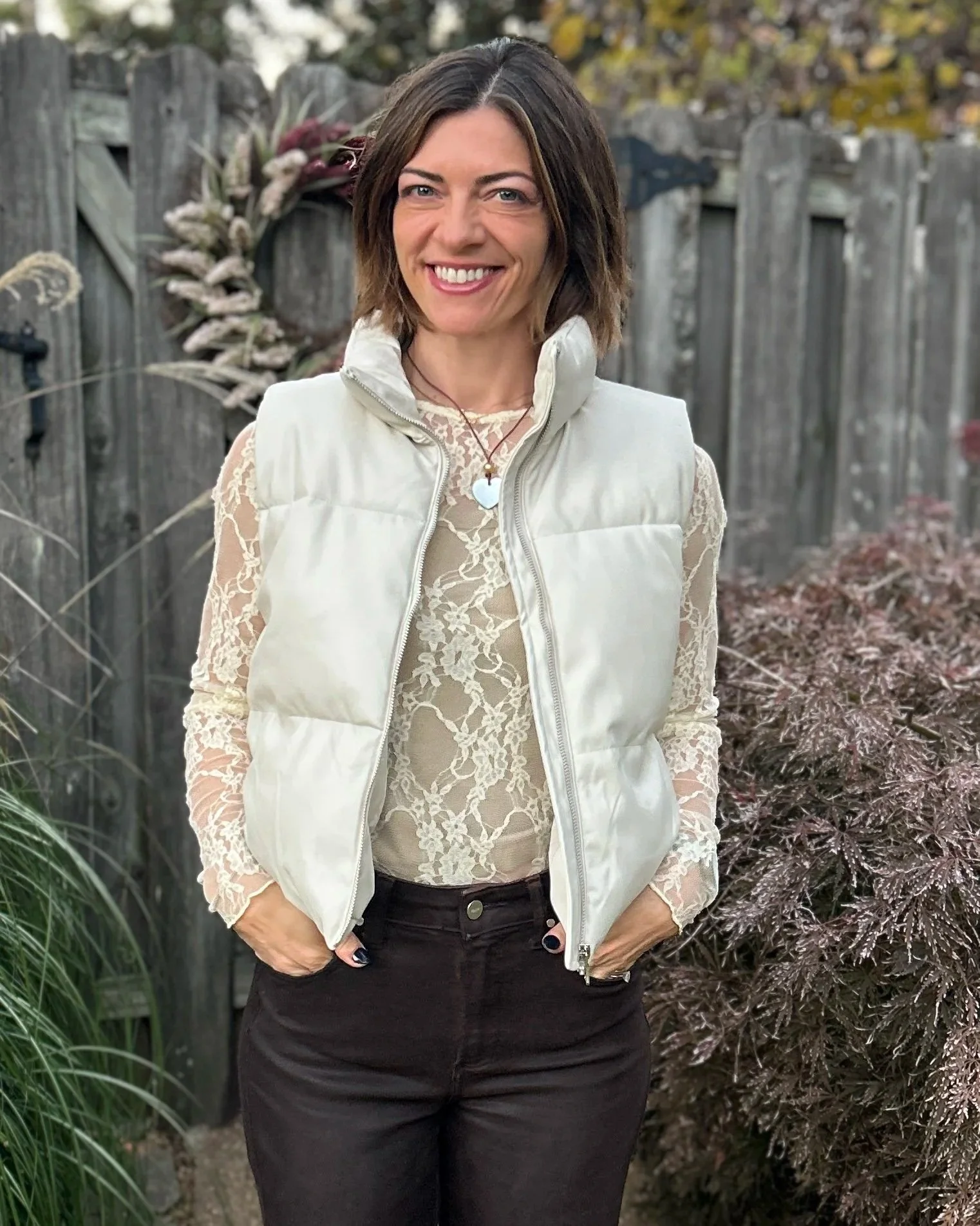 A woman with shoulder-length brown hair smiling outdoors, wearing a cream-colored puffer vest over a beige lace blouse, black pants, and a necklace with a heart-shaped pendant, standing in front of a wooden fence surrounded by foliage.