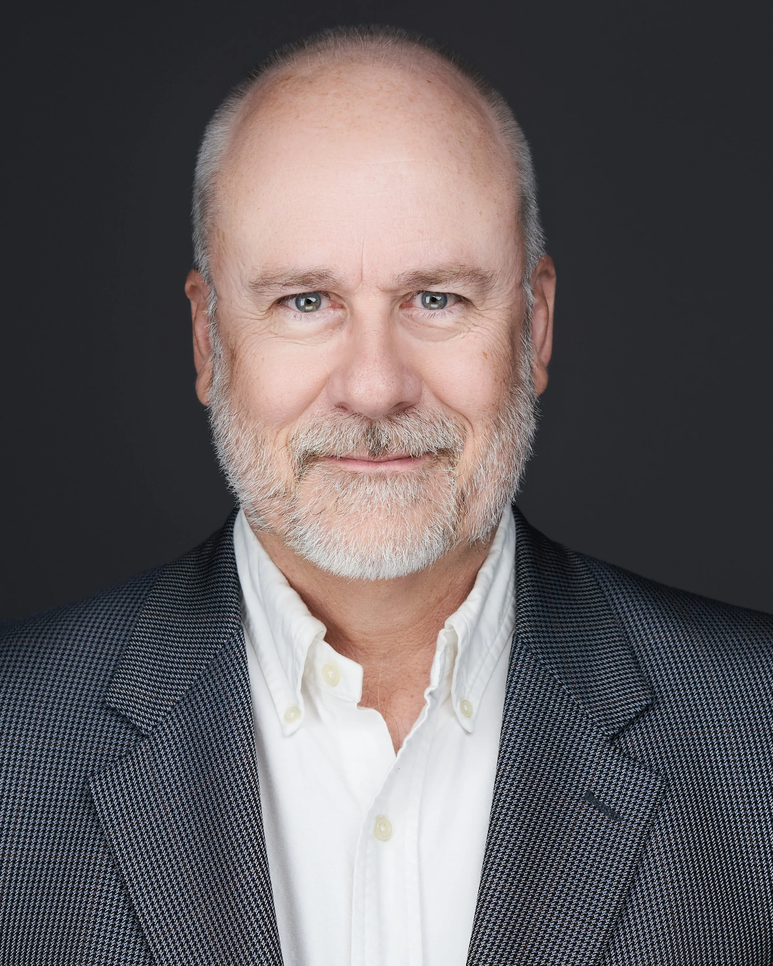 A middle-aged man with a beard and mustache smiling, wearing a white shirt and a patterned grey blazer, against a dark background.
