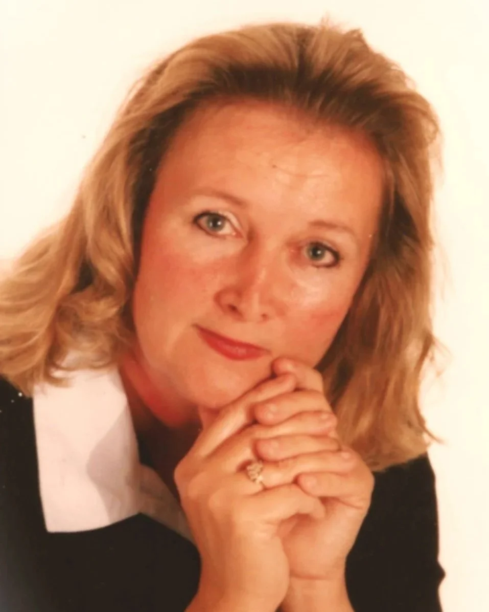 A woman with blonde hair, wearing a white collared shirt under a black top, resting her chin on her clasped hands and looking at the camera with a slight smile.