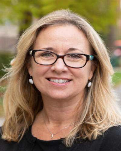 A smiling woman with blonde hair, glasses, and earrings outdoors.