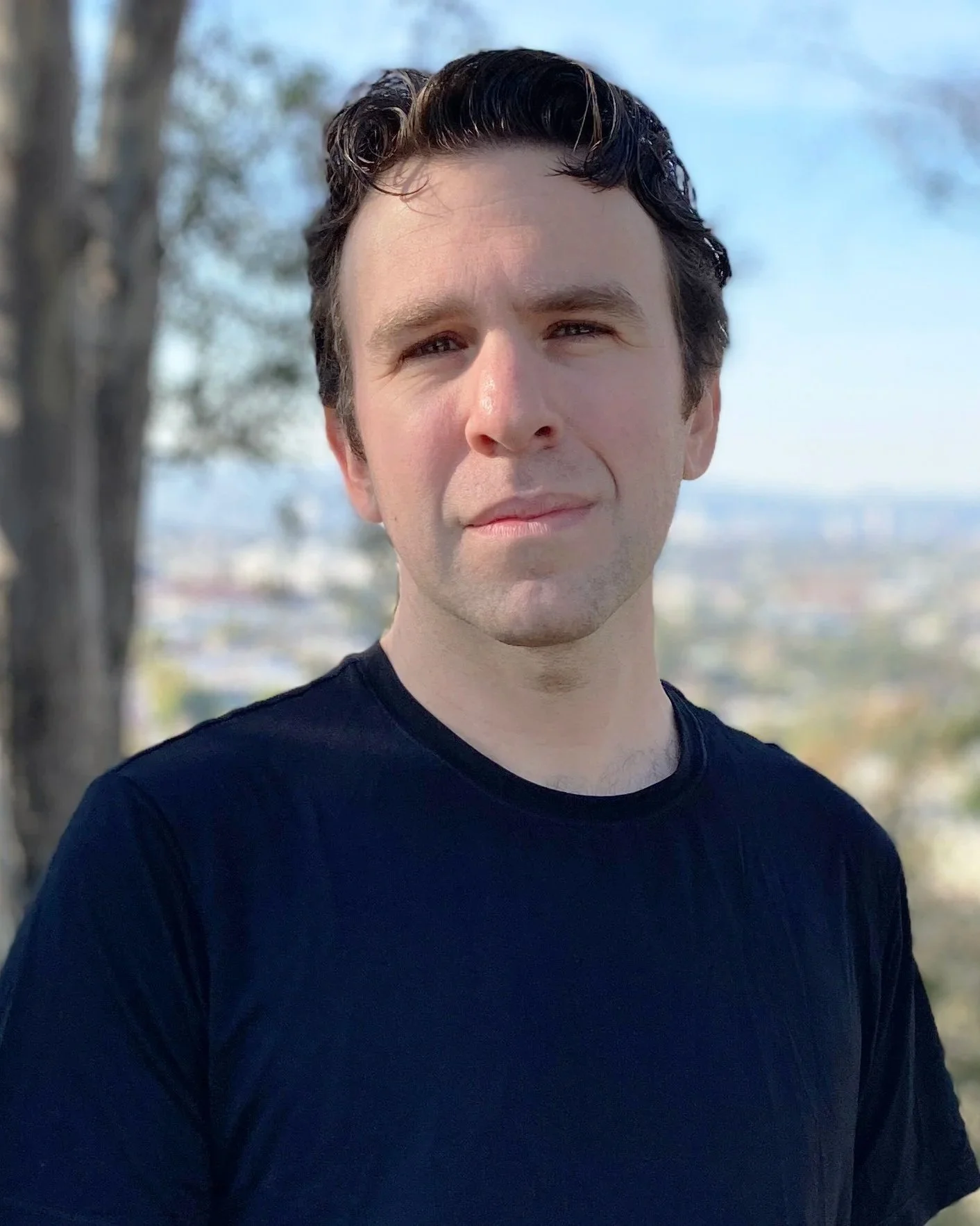 A young man with dark, wavy hair, wearing a black shirt, standing outdoors with blurred trees and cityscape in the background.