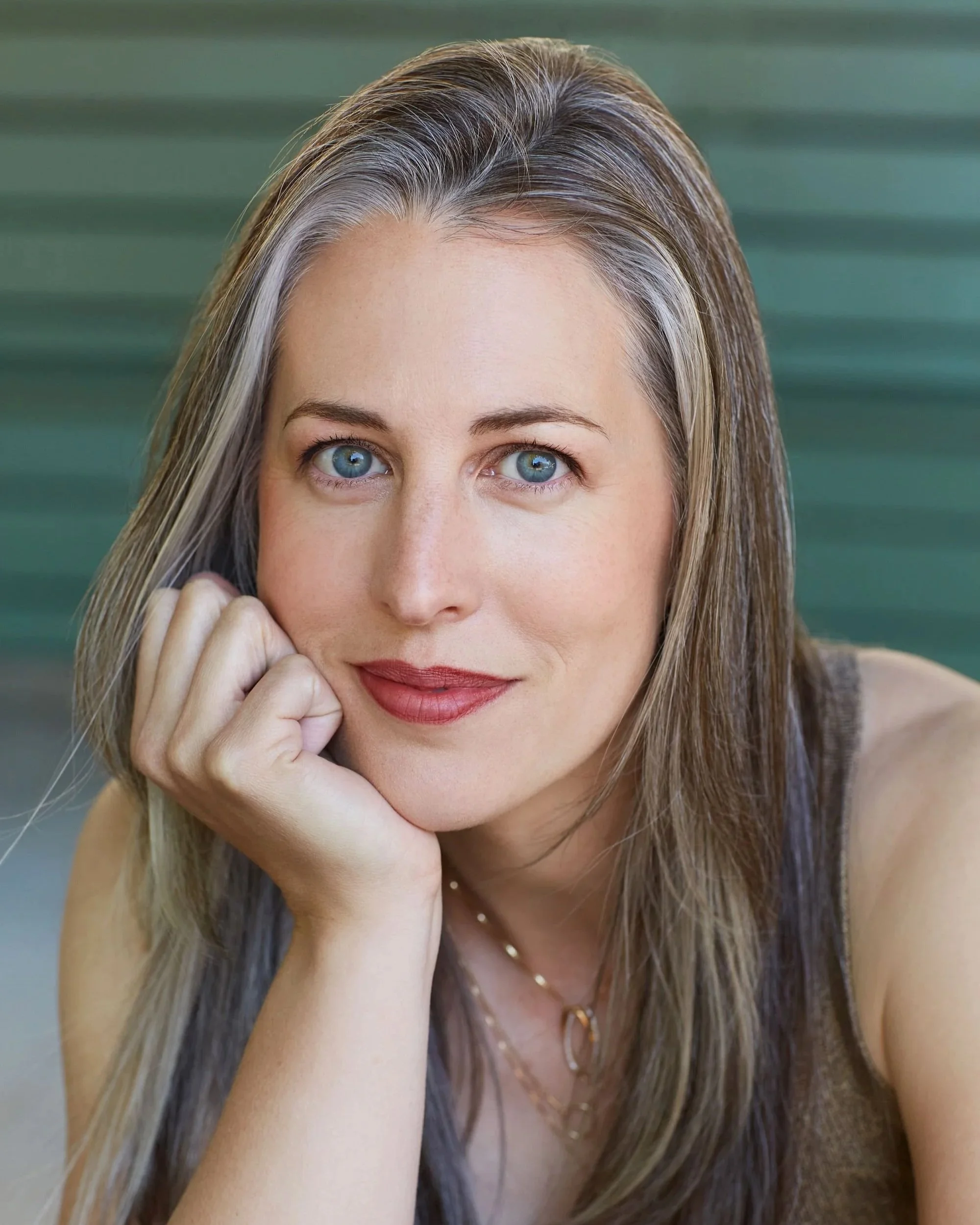 A woman with blue eyes and light brown hair resting her chin on her hand, smiling gently.