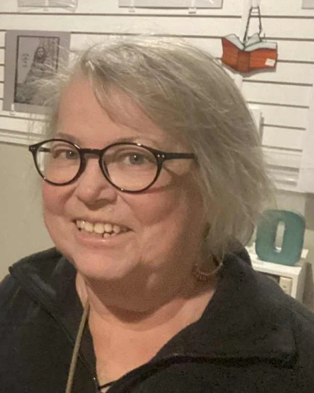 A woman with light-colored hair, glasses, and a black jacket smiling indoors. Background includes a window with blinds, a small decorative ladder, and a teal-colored decorative object.