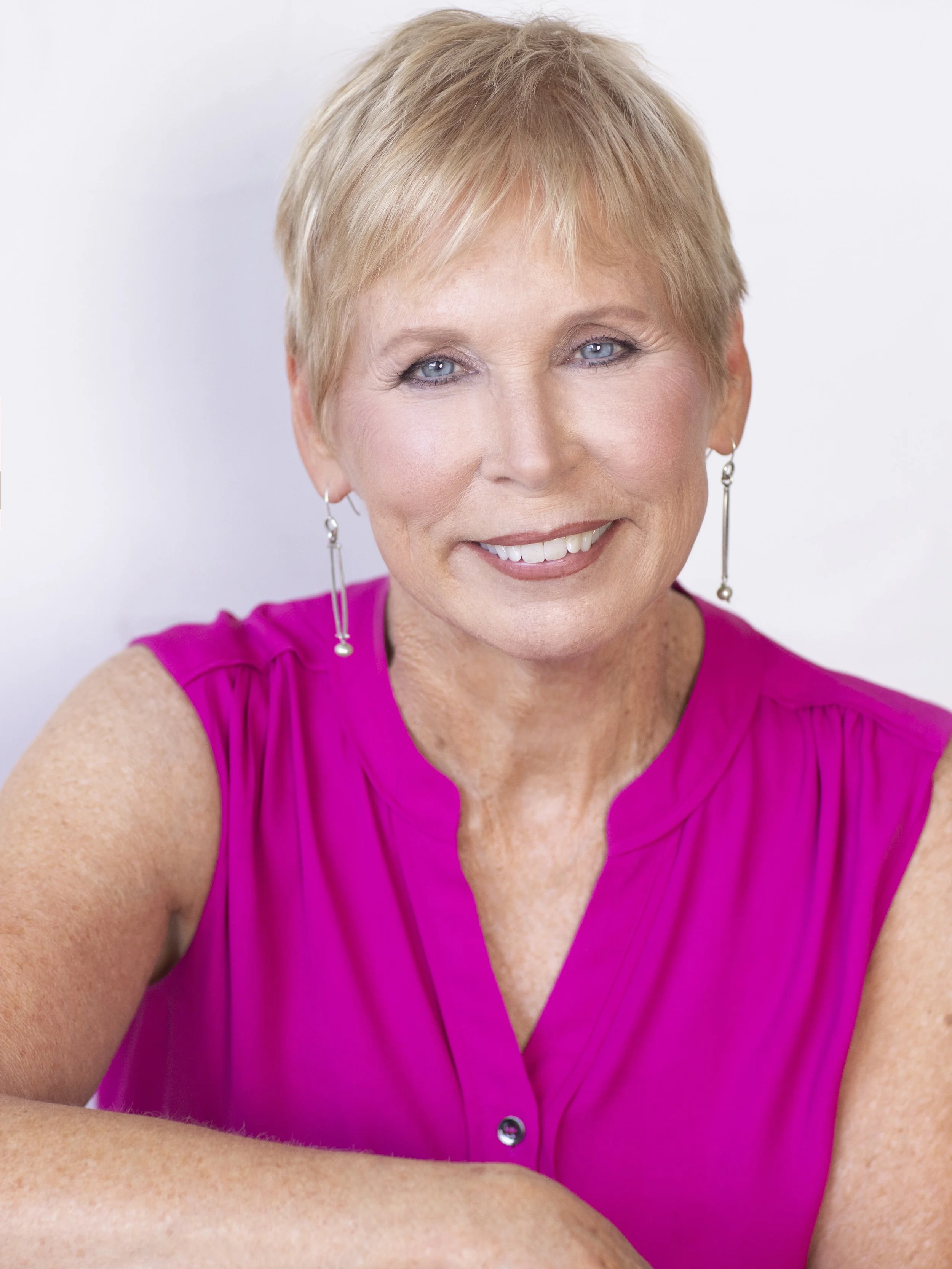 A smiling woman with short blonde hair, wearing a bright pink sleeveless blouse and silver earrings, against a plain white background.
