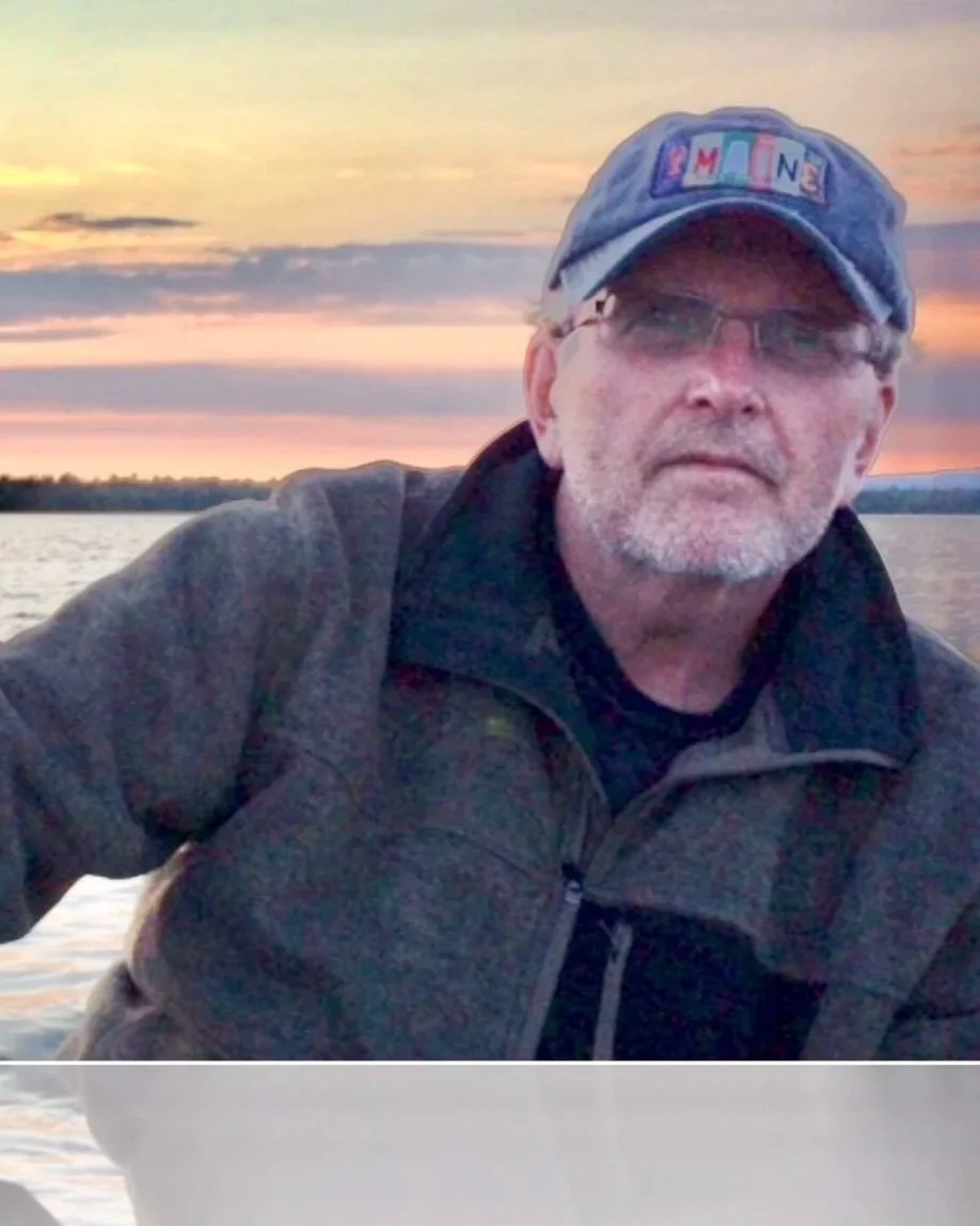 A man with glasses and a gray beard wearing a cap with colorful letters spelling 'MINE' and a gray jacket, sitting outdoors near a body of water during sunset.