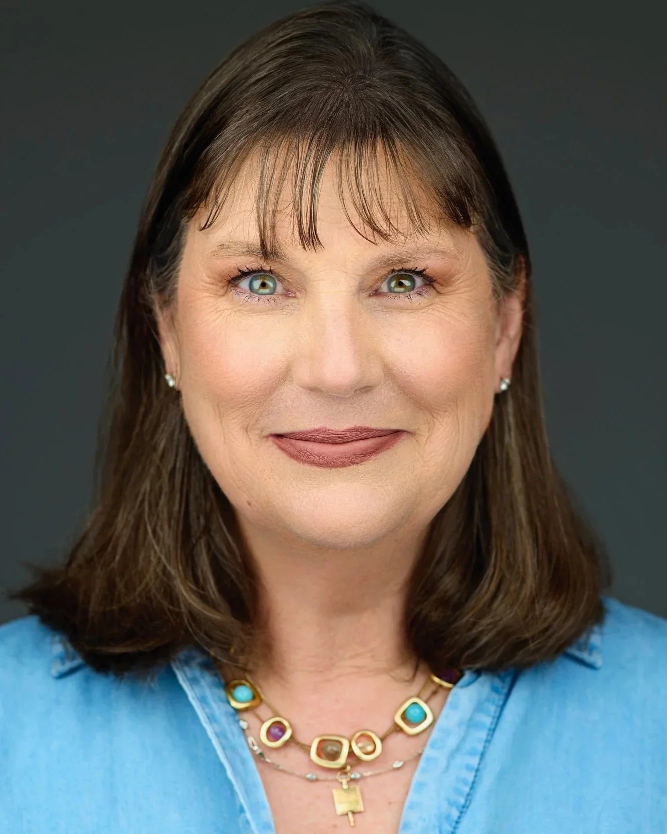 Close-up of a middle-aged woman with brown hair, green eyes, wearing a blue top and a colorful necklace, smiling against a dark background.
