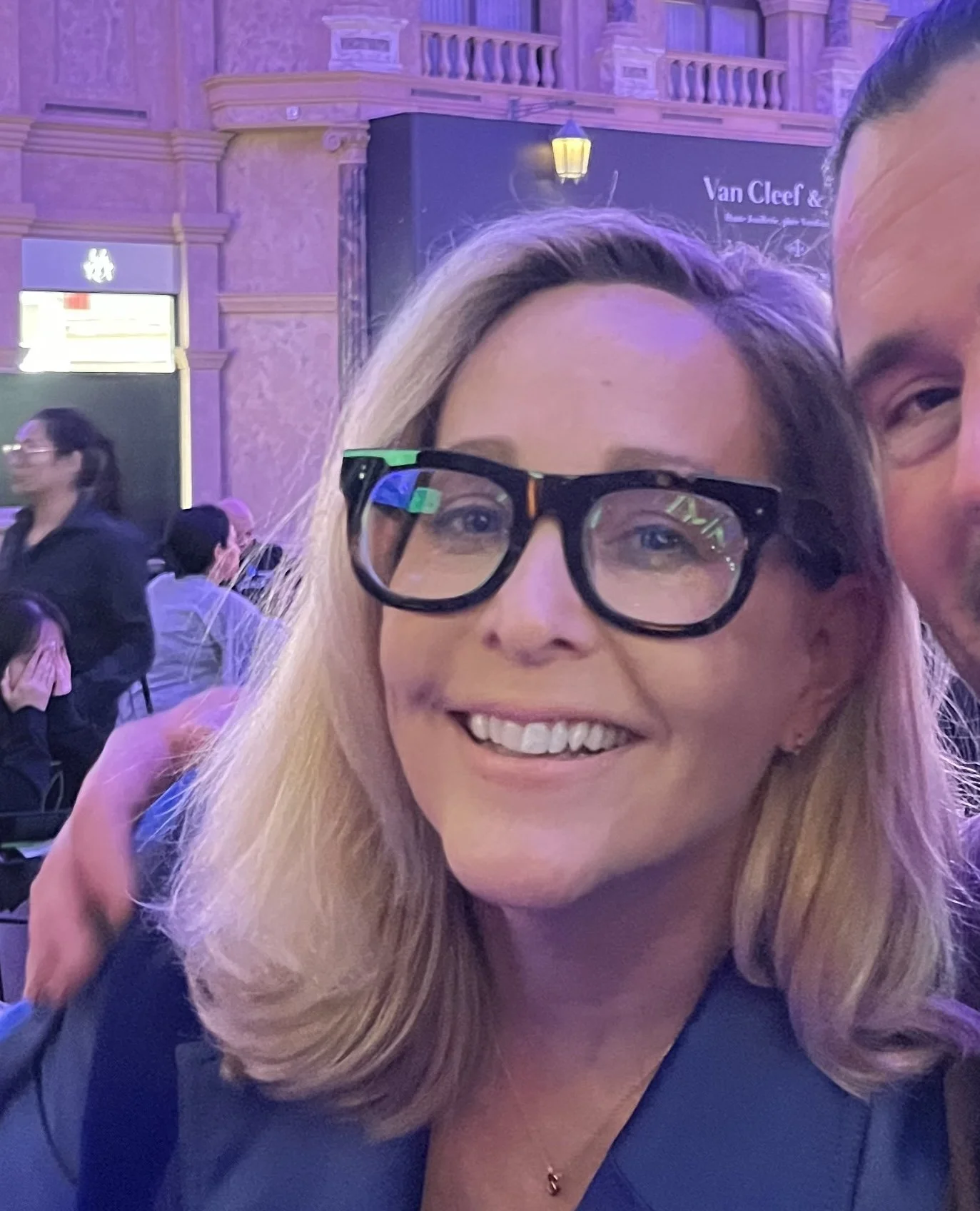 A smiling woman with blonde hair and glasses, wearing a blue blazer, taking a selfie in an indoor event space with purple lighting and people in the background.